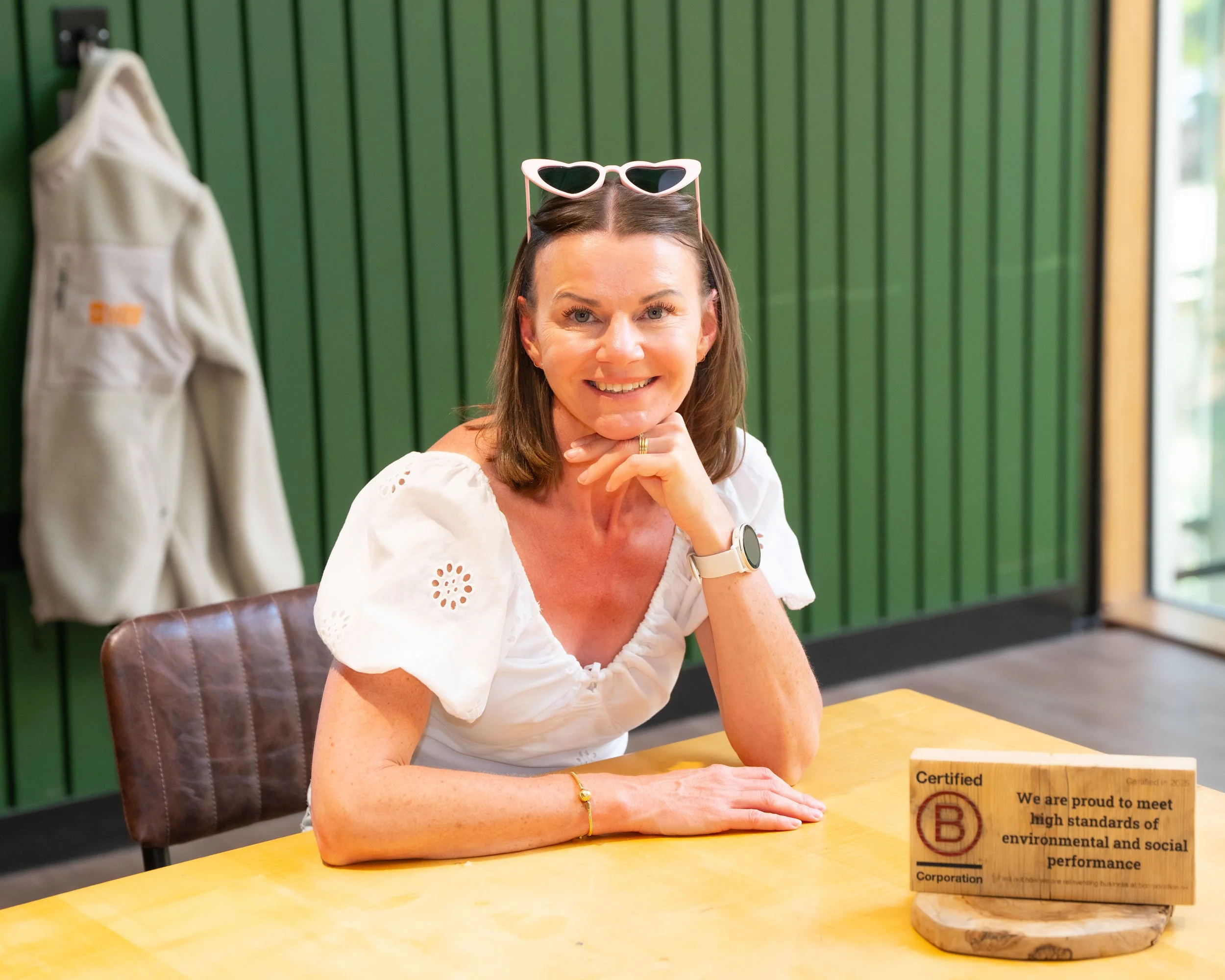 A smiling woman with brown hair, wearing white sunglasses on her head, is sitting at a wooden table with a green paneled wall behind her. She is resting her chin on her right hand, and there is a small wooden sign with text on the table.