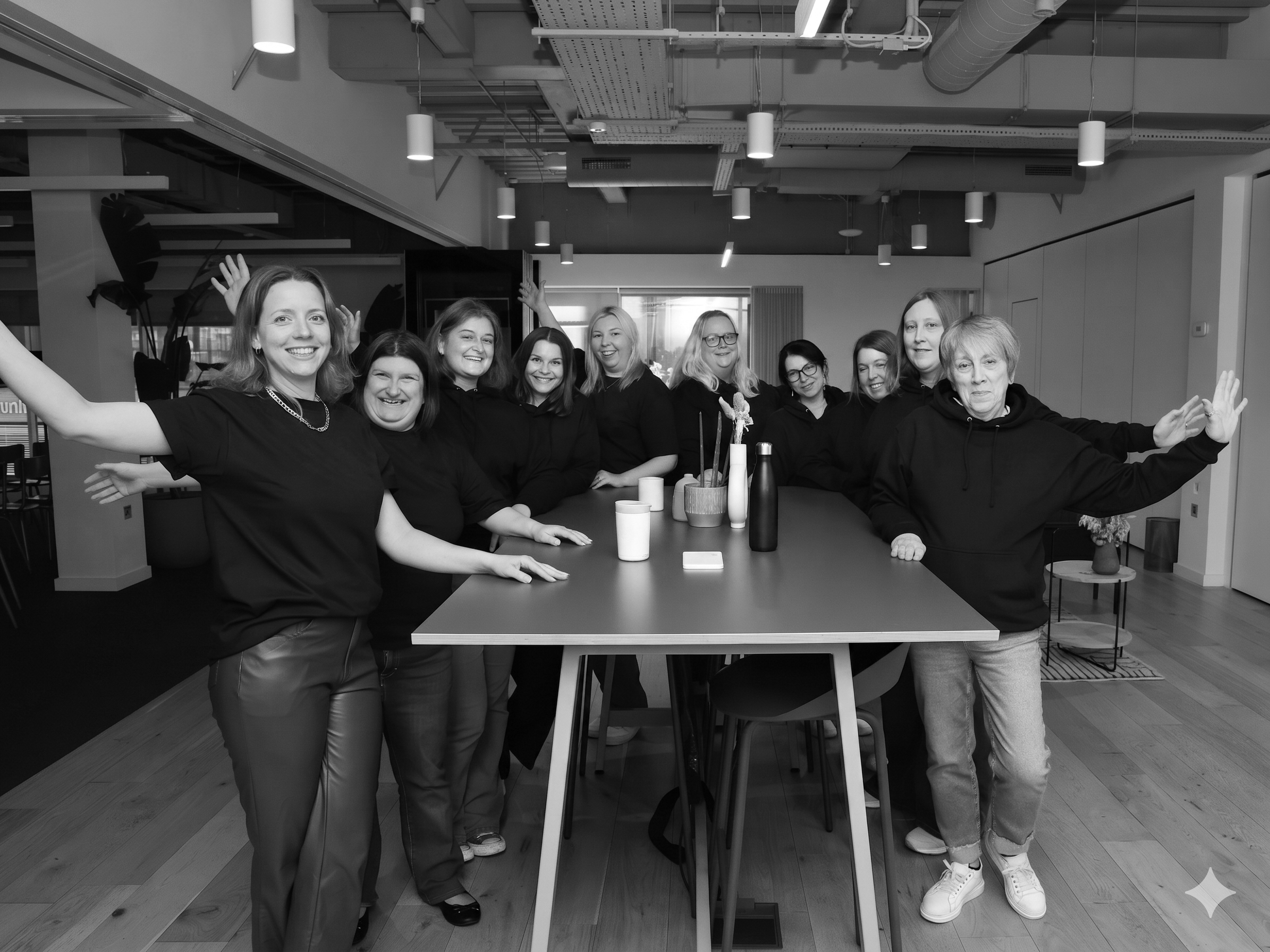 Group of nine women smiling and posing with arms raised inside a modern office or conference space with ceiling lights and large windows.