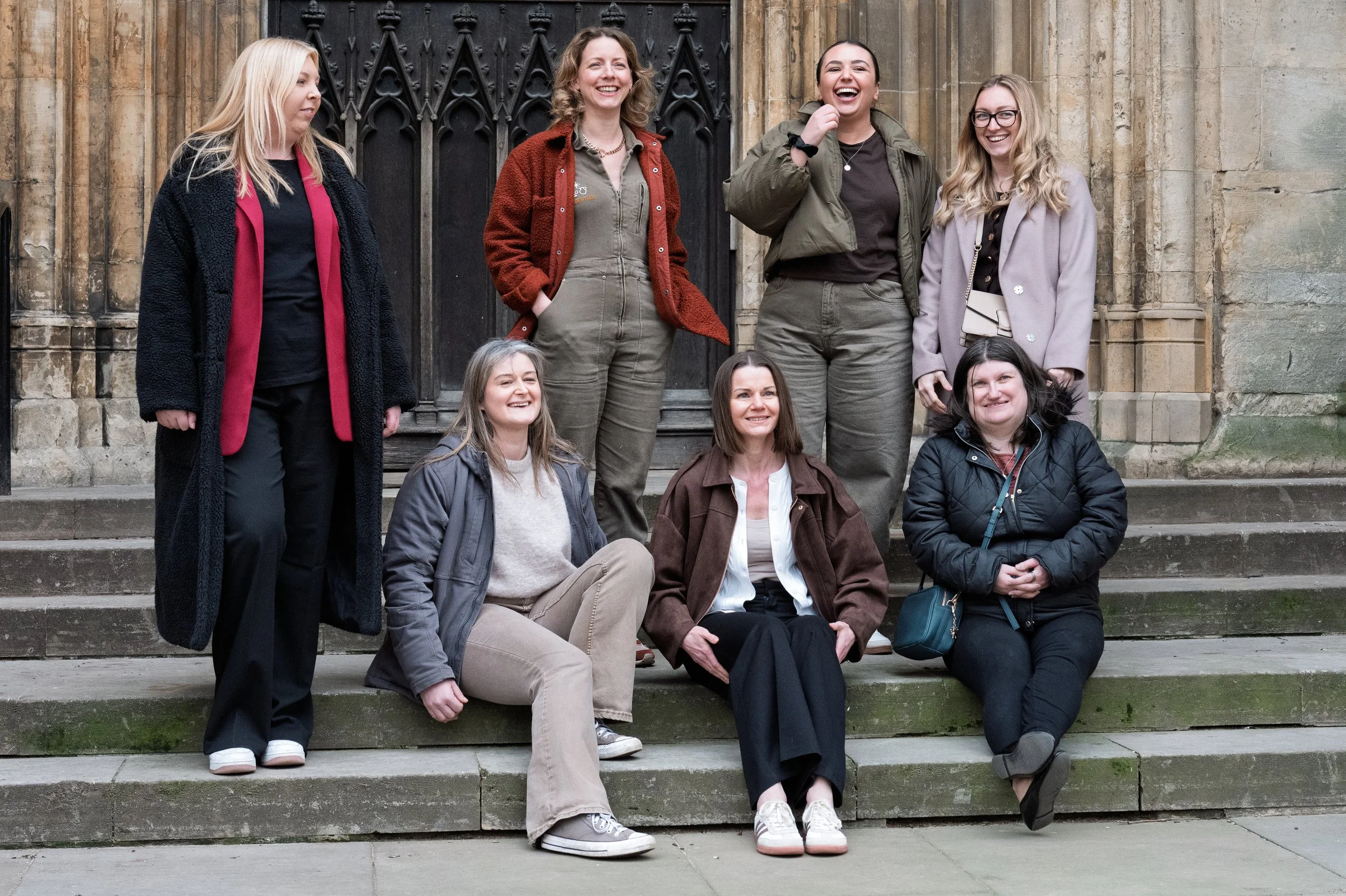Group of seven women outside on stone steps, some sitting and some standing, smiling and chatting, near a historic building with gothic architecture elements.