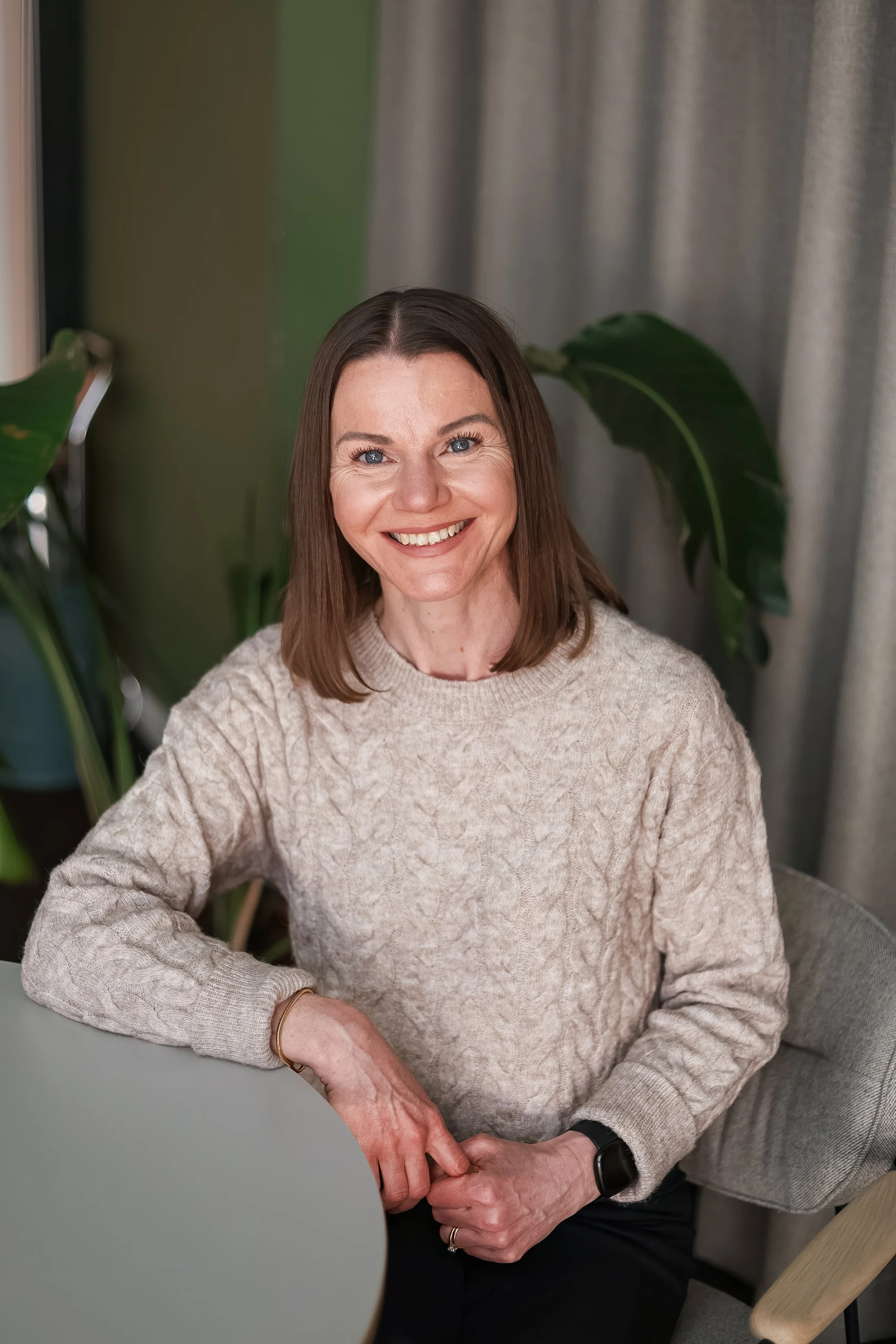 A woman with shoulder-length brown hair, blue eyes, and a warm smile, wearing a beige cable knit sweater and a black watch, sitting at a table in a cozy indoor setting with green and gray walls and large leafy houseplants nearby.