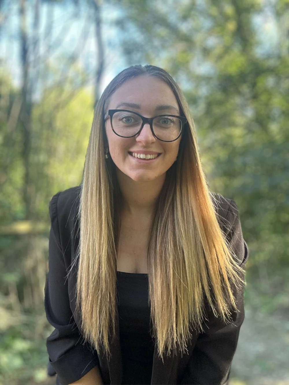 A young woman with long straight blonde hair and glasses is smiling outdoors with a blurred green forest background.