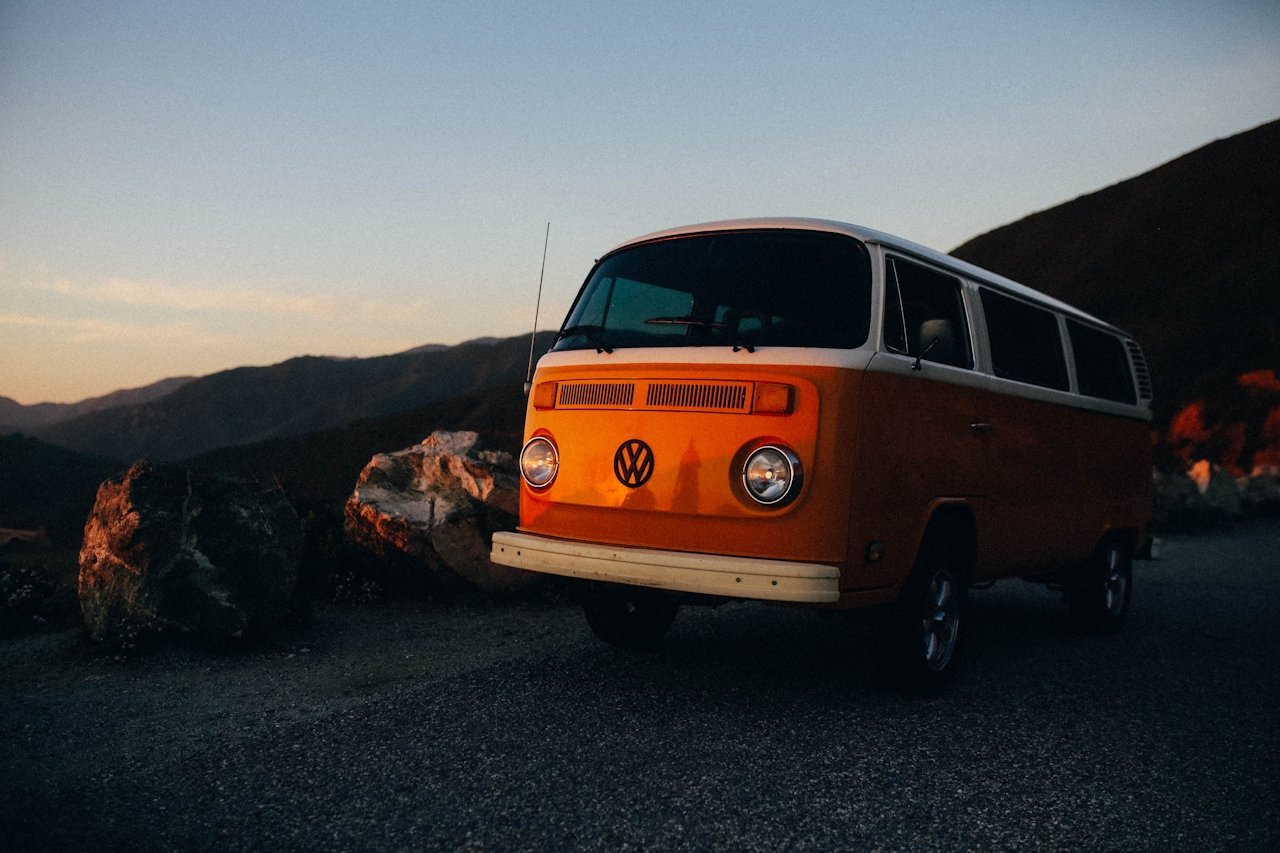 A vintage orange Volkswagen van parked on a mountain road during sunset, with rocks and mountain hills in the background.