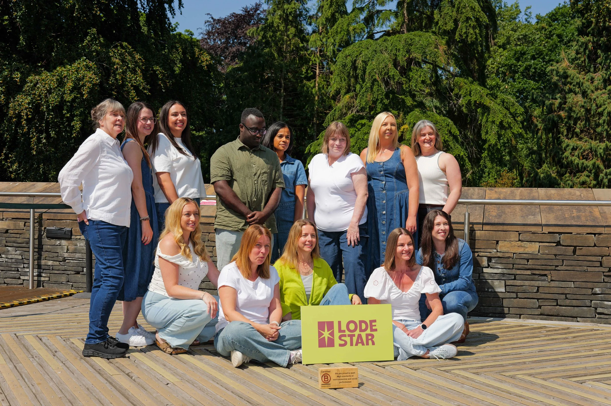 Group of women and men posing outdoors for a photo on a wooden platform, with lush green trees in the background. Some are standing, some are sitting, and there is a sign that reads 'LODE STAR' in front of them.