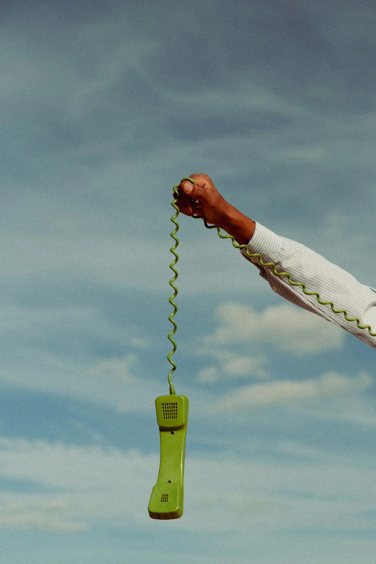 Person holding a vintage green telephone receiver connected to a wire hanging in the air against a cloudy sky.