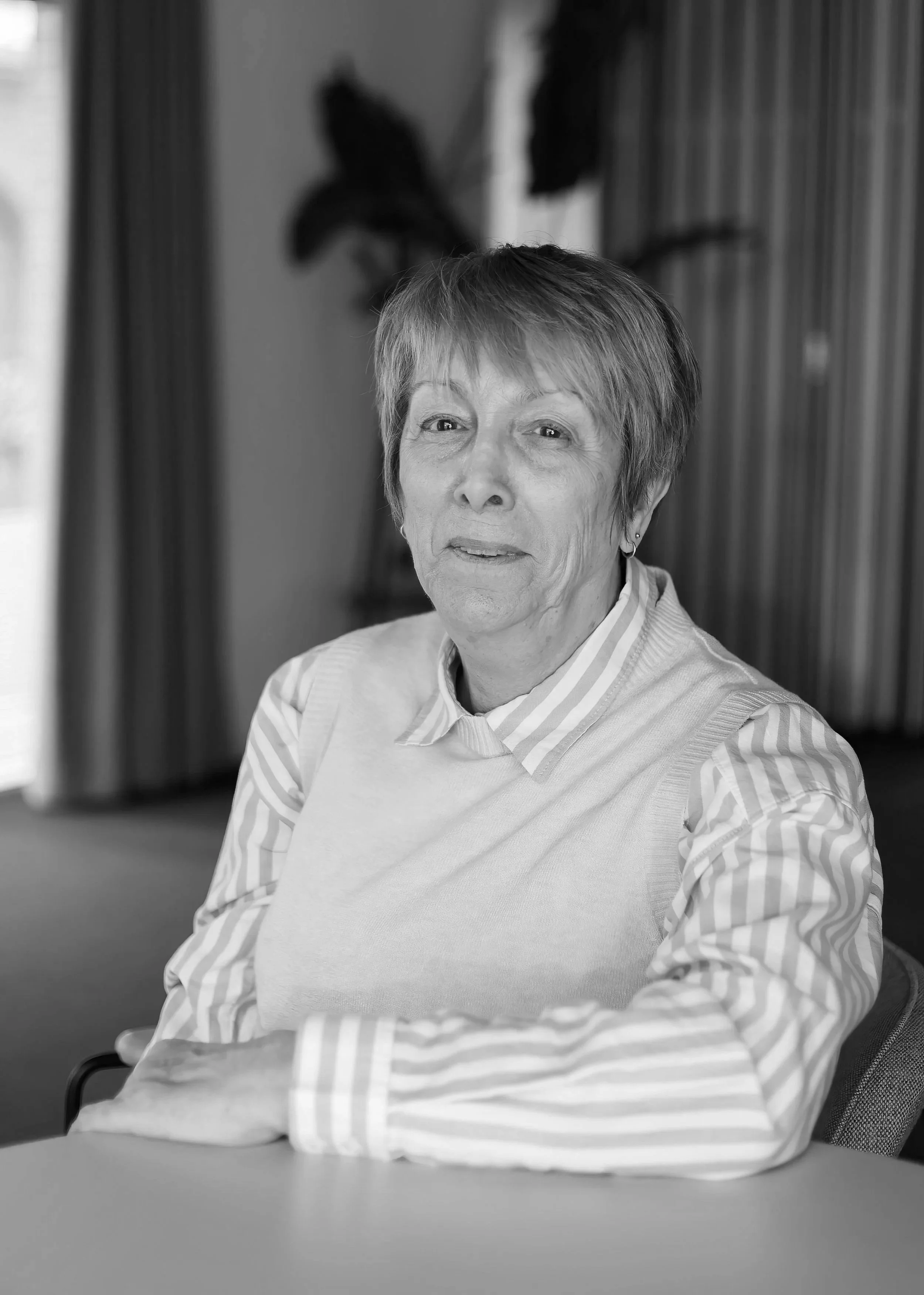 A senior woman with short hair and earrings sitting at a desk, looking slightly to the side, in black and white.