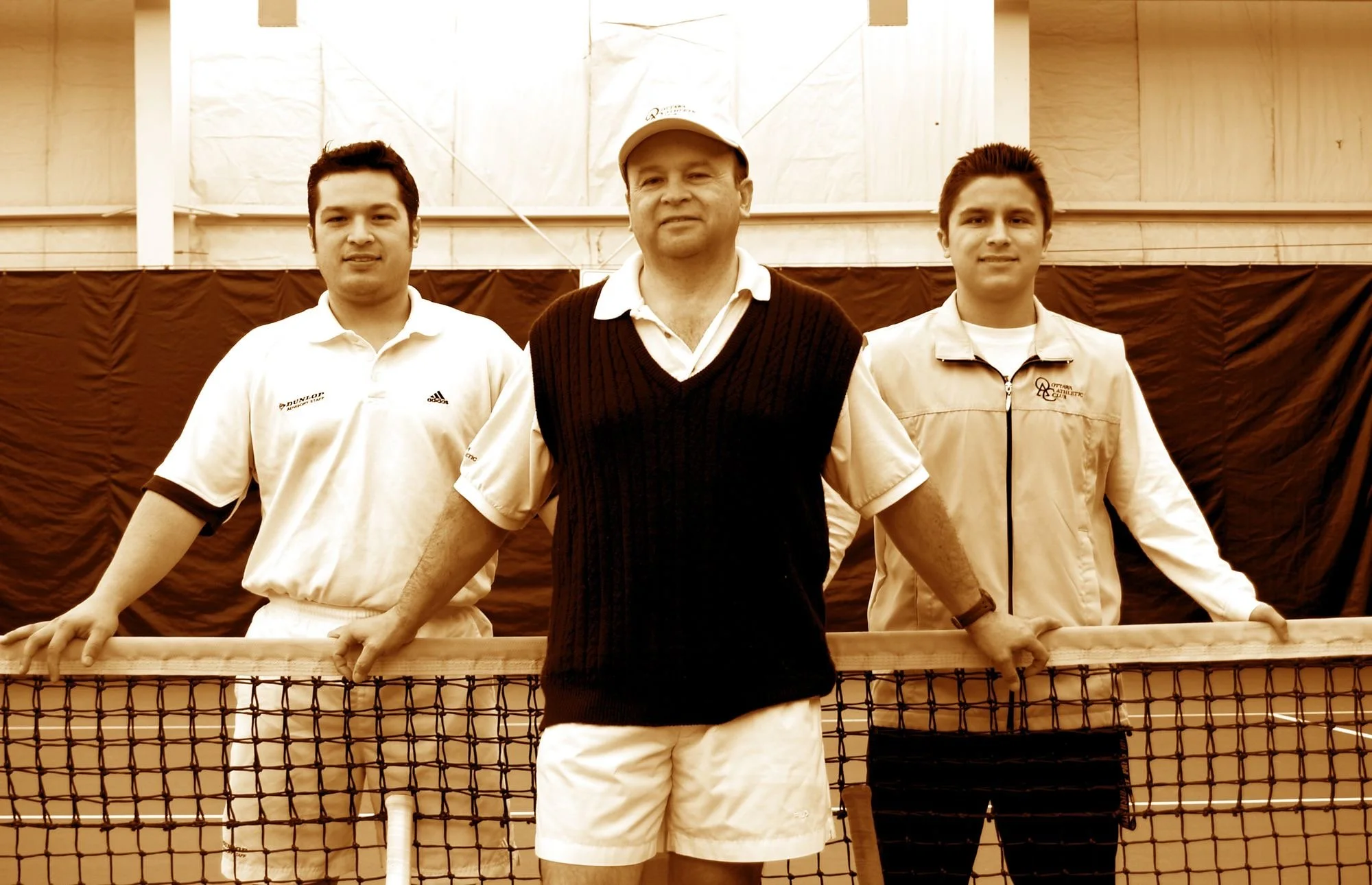 Three men standing behind a tennis net indoors. The man in the middle is wearing a baseball cap and a sweater vest, with two young men on either side wearing tennis attire. They are all holding onto the tennis net.
