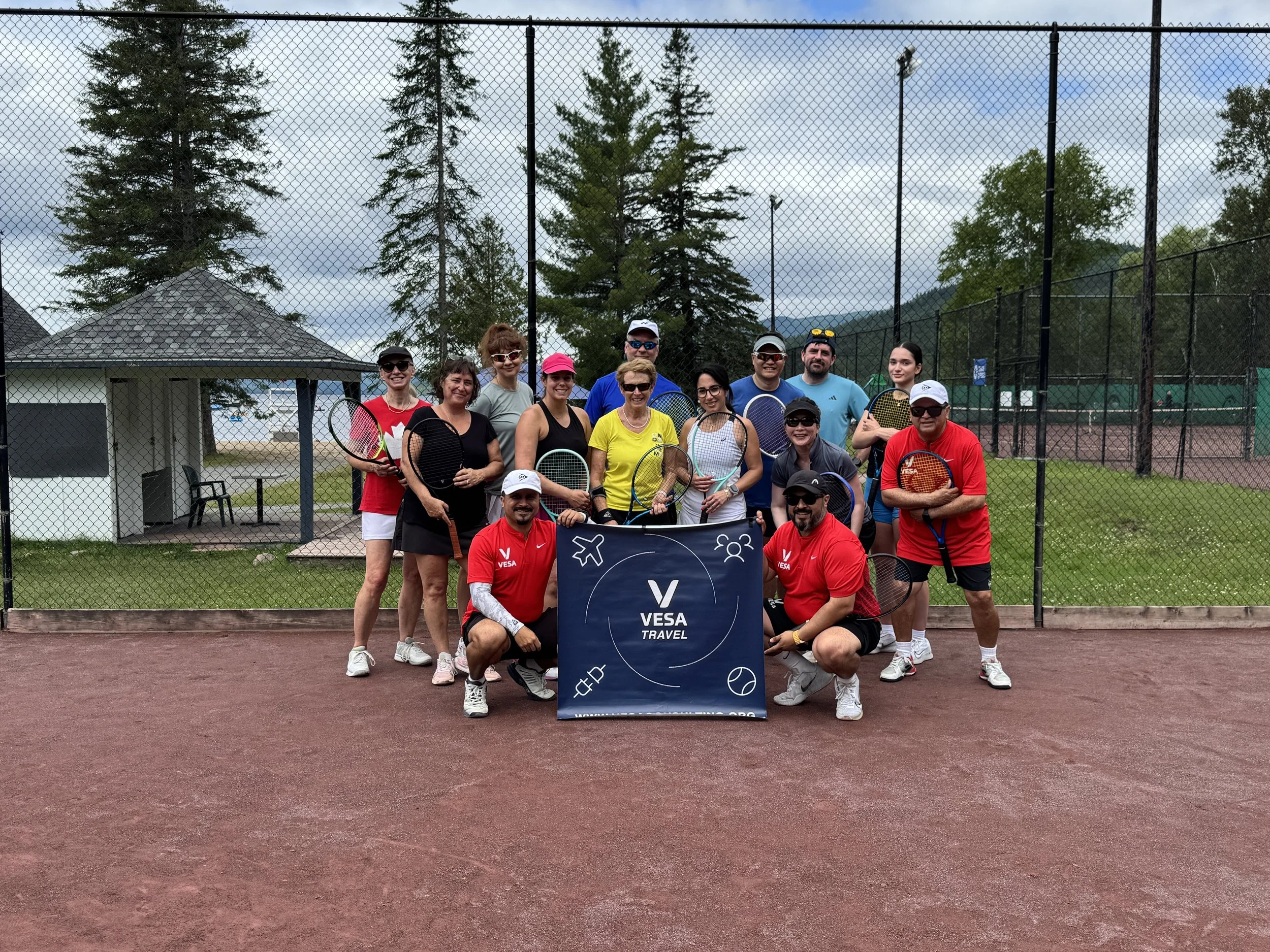 A group of people posing on a tennis court behind a chain-link fence. They are holding tennis rackets and wearing athletic clothing, with some wearing sunglasses. The background includes tall trees, a small building, and a cloudy sky.
