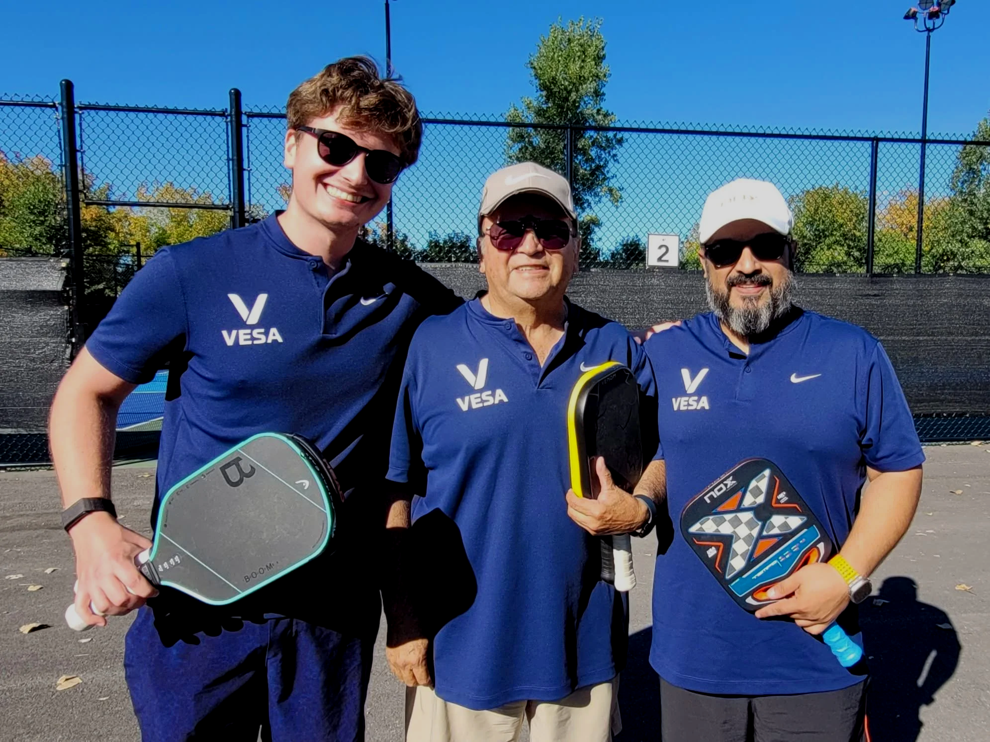 Three men wearing sunglasses and blue shirts with 'VESA' logos, holding padel rackets, standing outdoors on a sunny day in front of a black sports fence with a tennis court and a sign with the number 2, trees, and a blue sky in the background.