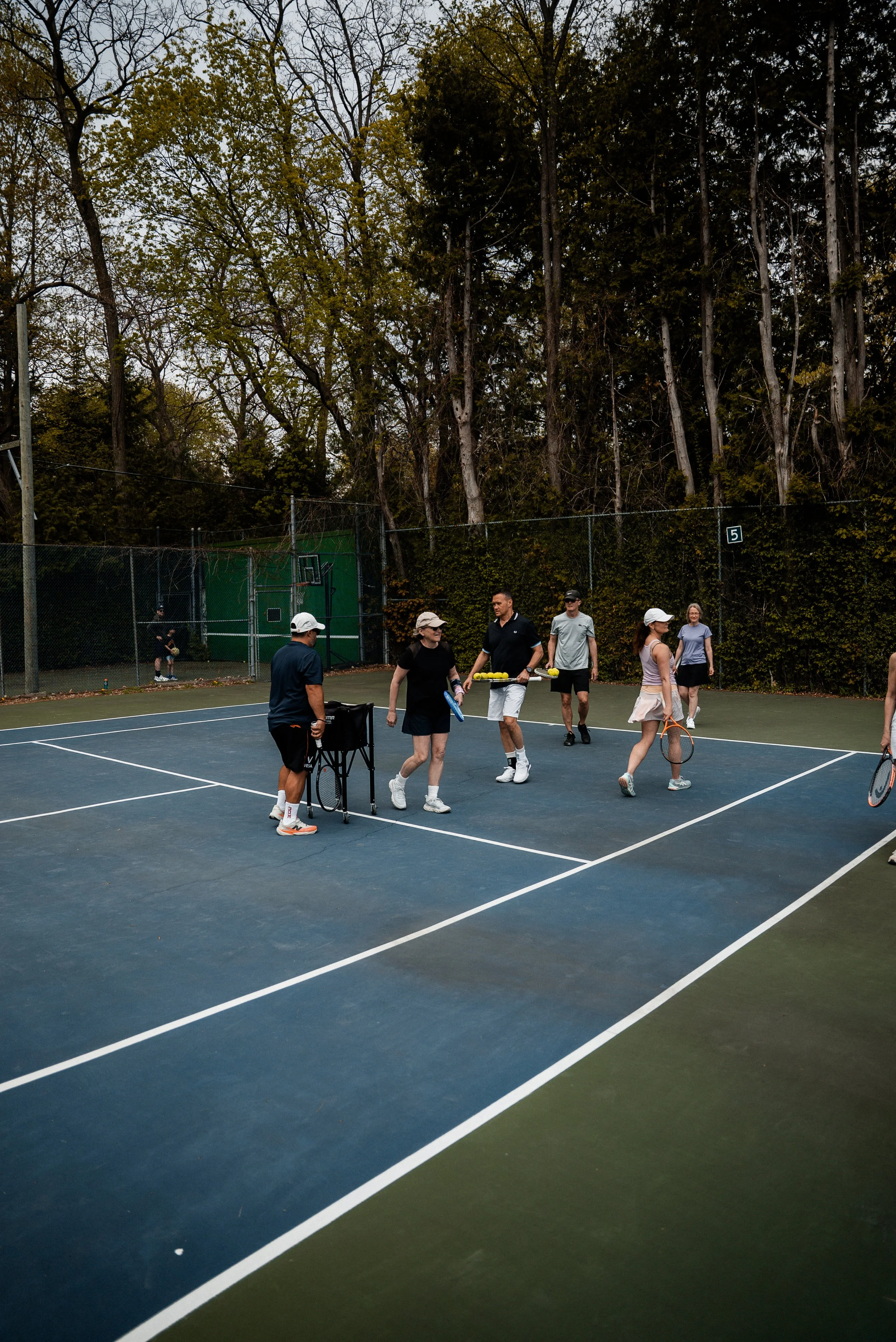 People on a tennis court, some holding tennis rackets, with a group of players in the center and others observing, surrounded by tall trees.