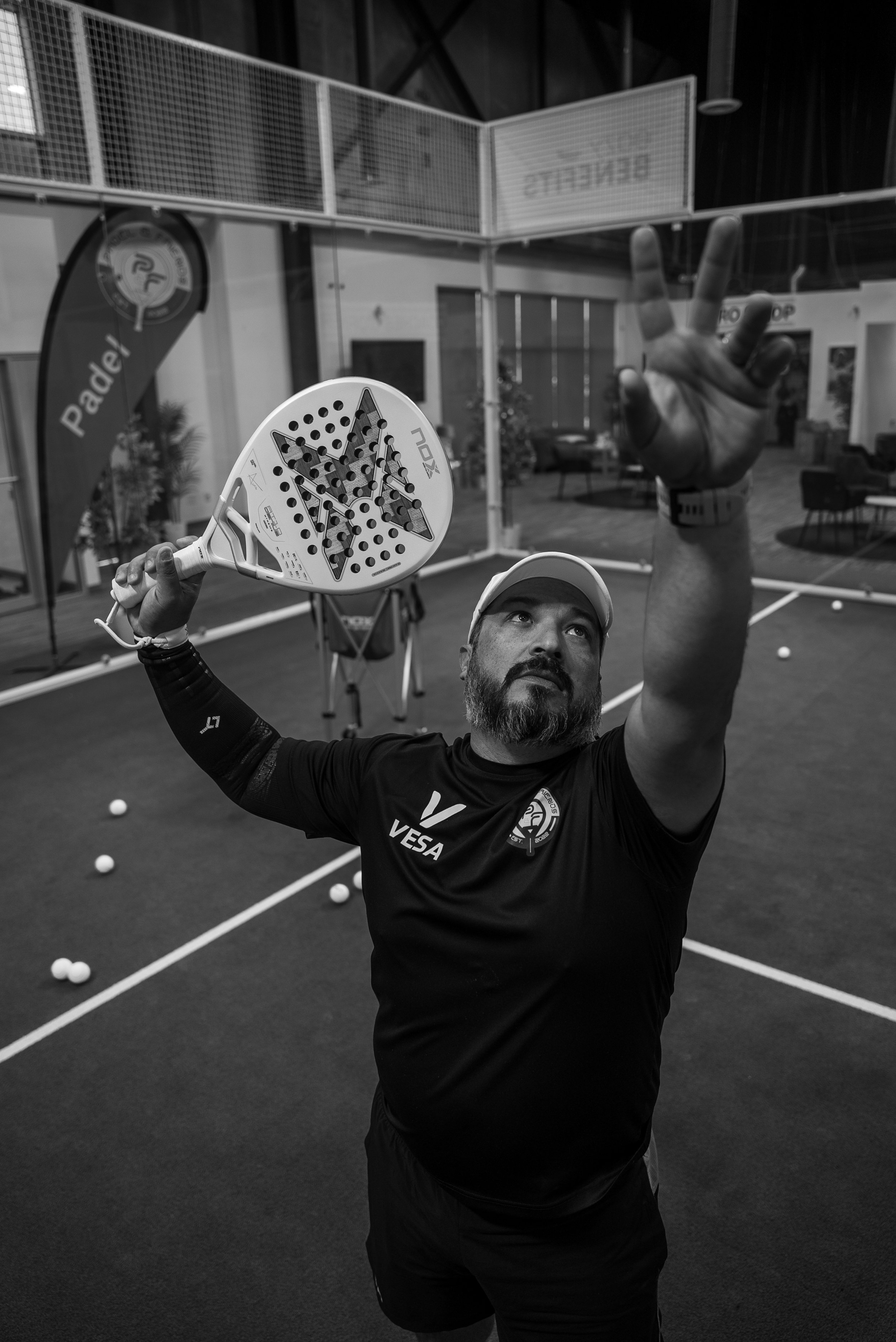 A man with a beard and wearing a white cap and a black t-shirt with logos, holding a padel racket and making a peace sign with his right hand inside an indoor padel court.