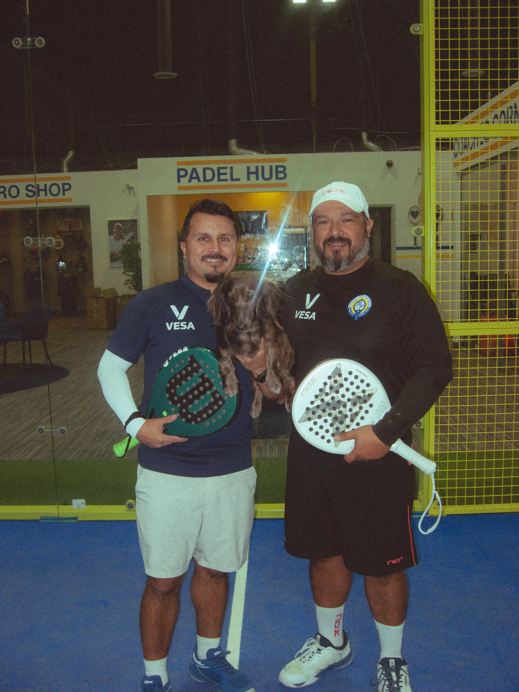Two men with athletic builds standing on an indoor pickleball court, holding paddles, smiling at the camera, with one man holding a small dog. Behind them, there is a sign that reads 'Padel Hub'.