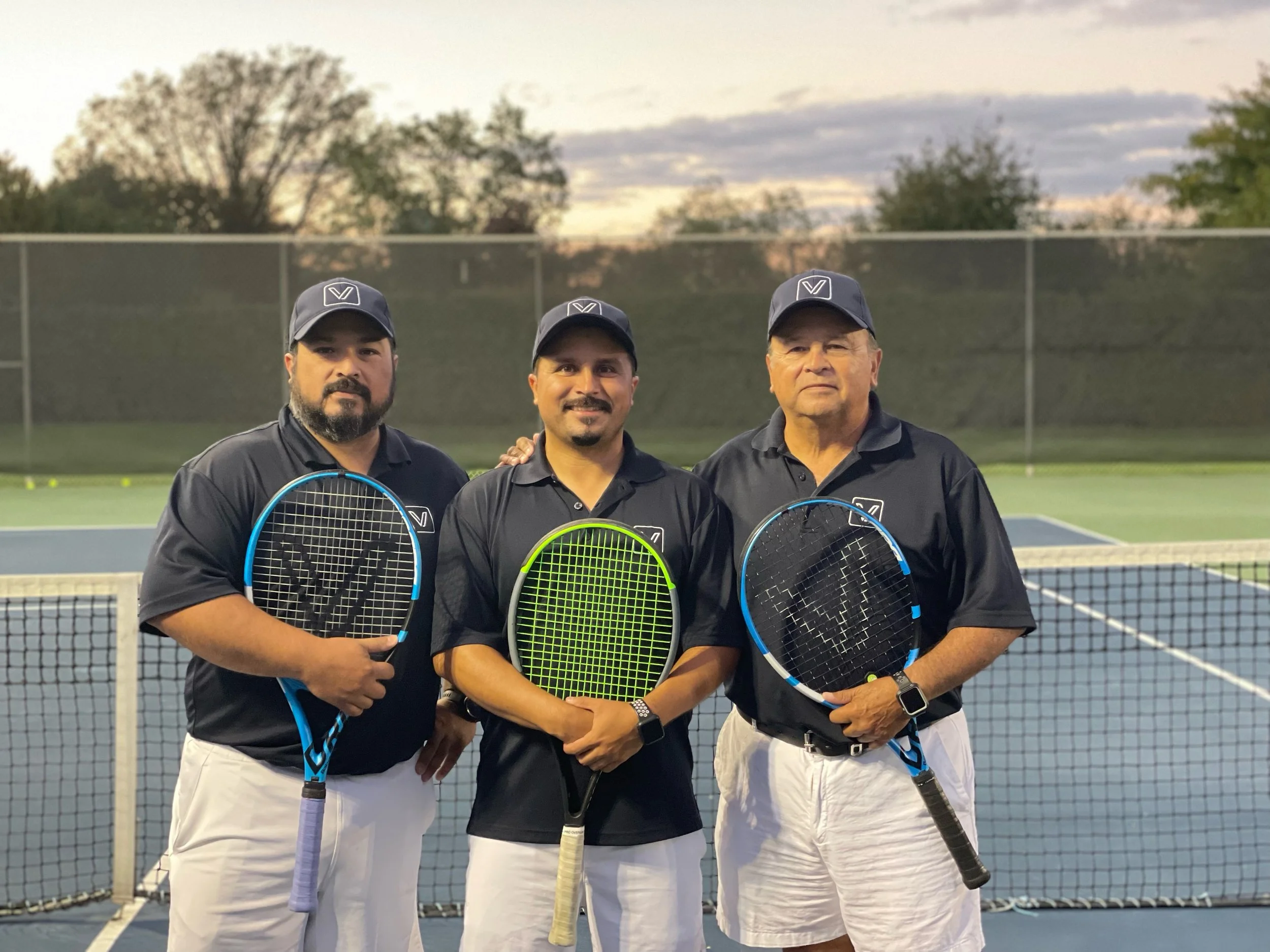 Three men in black shirts and white pants standing on a tennis court with tennis rackets, with a fence and trees in the background during dusk.