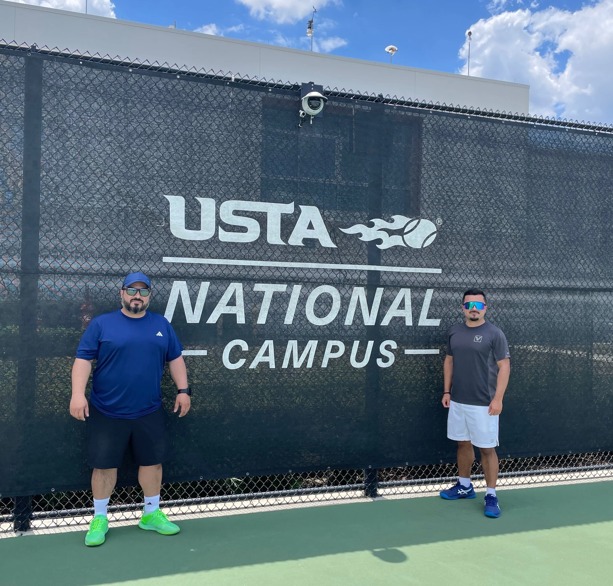 Two men standing on a tennis court in front of a large USTA National Campus sign, wearing athletic clothes and sunglasses, with a chain-link fence and a blue sky with clouds in the background.