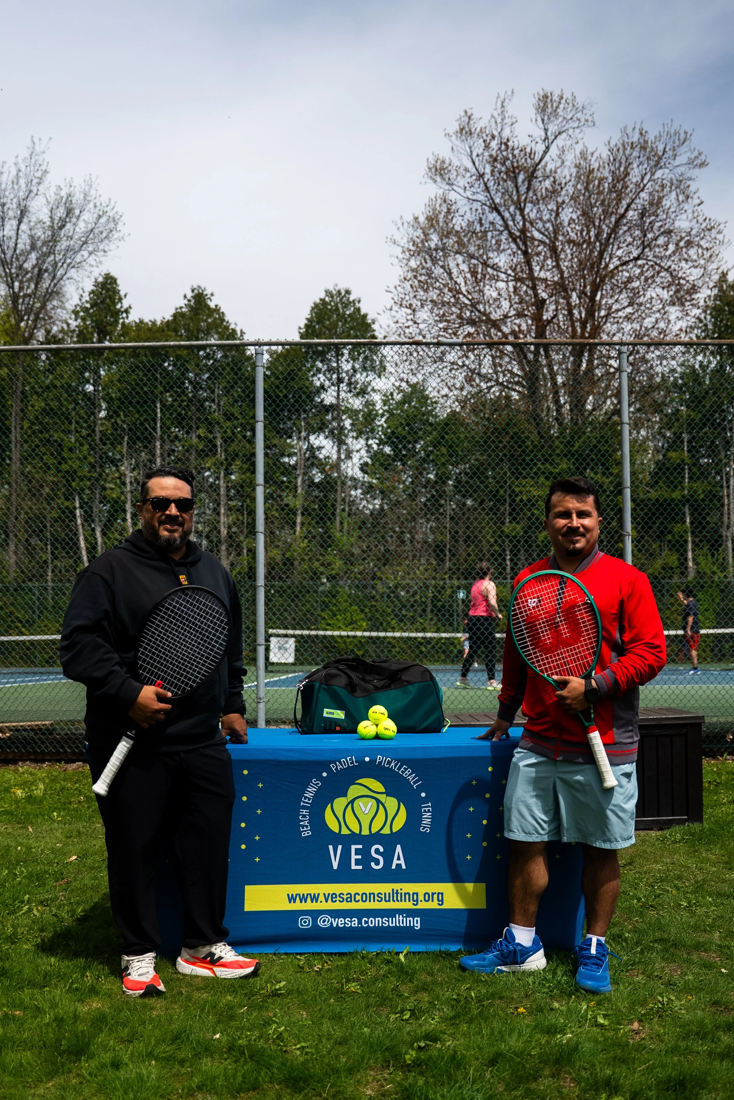 Two men standing outdoors in front of a table with tennis balls and a VESA sign, holding tennis rackets, on a tennis court with trees and a chain-link fence in the background.