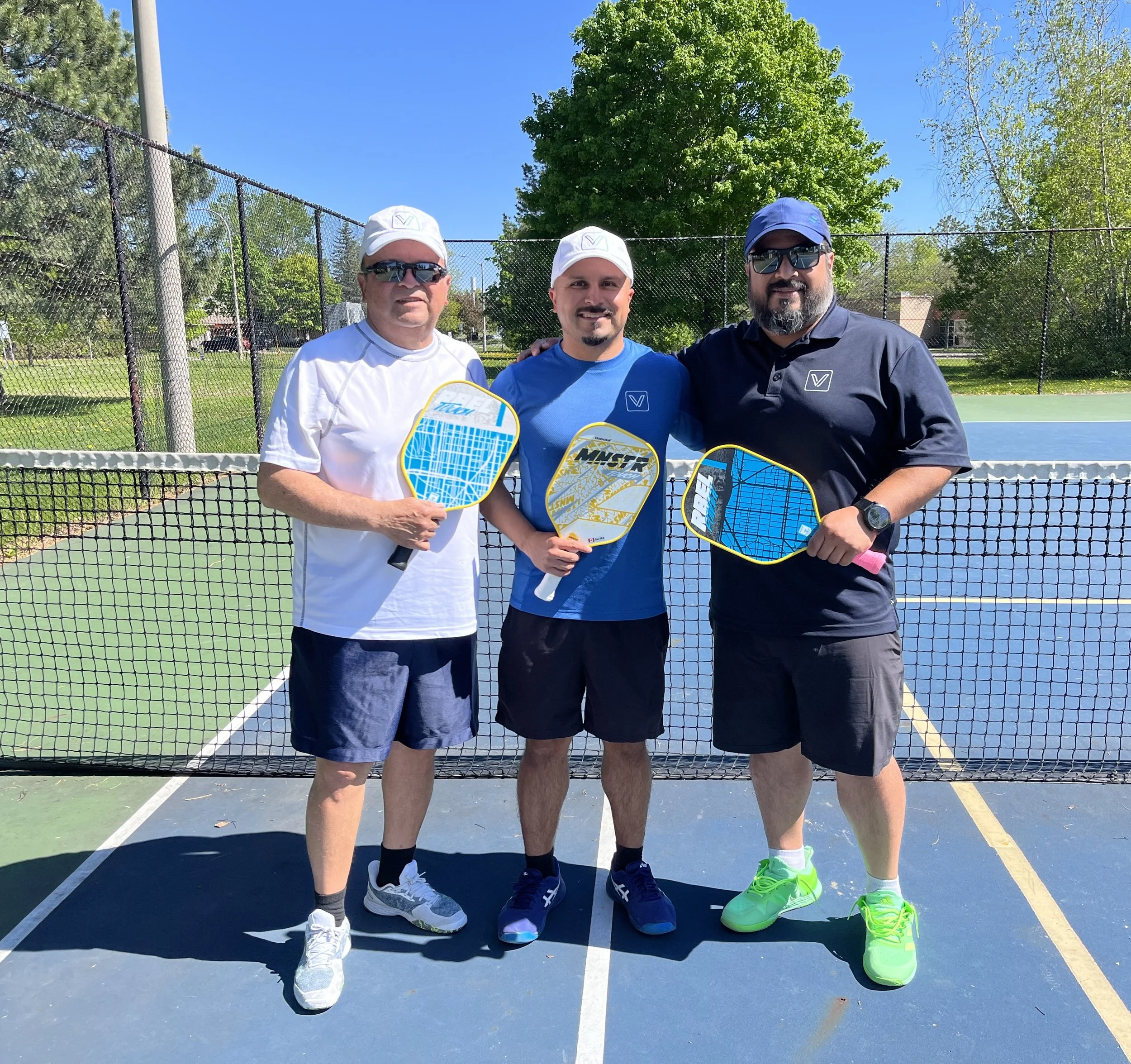 Three men standing together on a tennis court, holding pickleball paddles. They are dressed in athletic clothing, wearing sunglasses and caps, with greenery and a blue sky in the background.