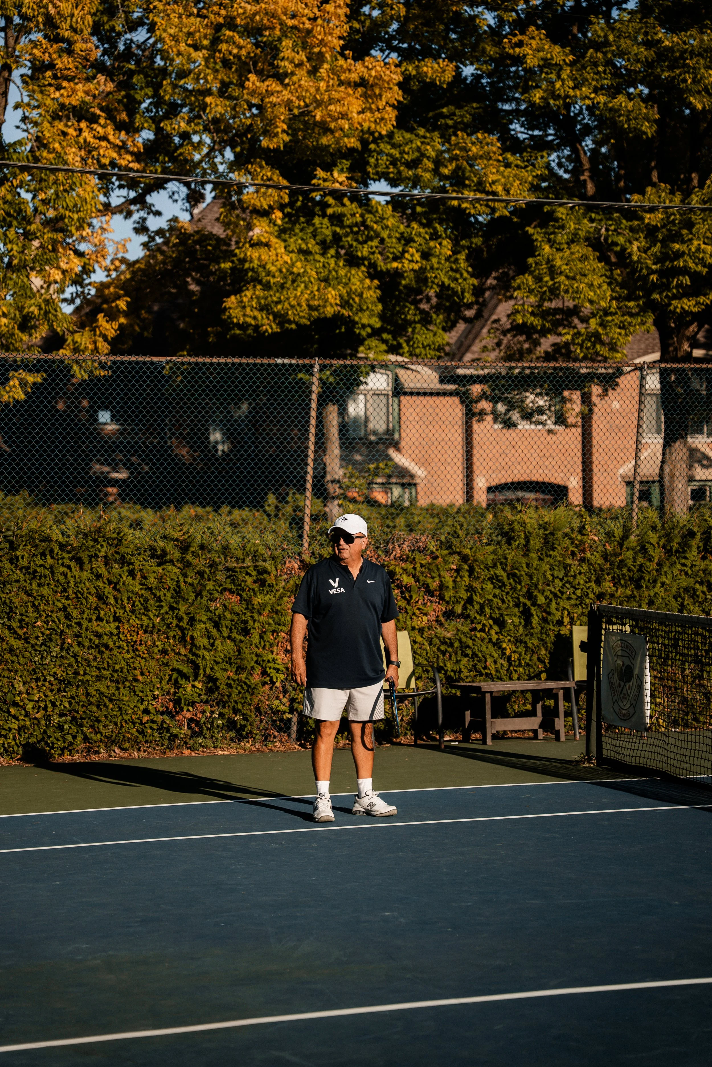 A man stands on a tennis court holding a tennis racket, wearing a black shirt, white shorts, and a white cap, with trees and houses in the background.