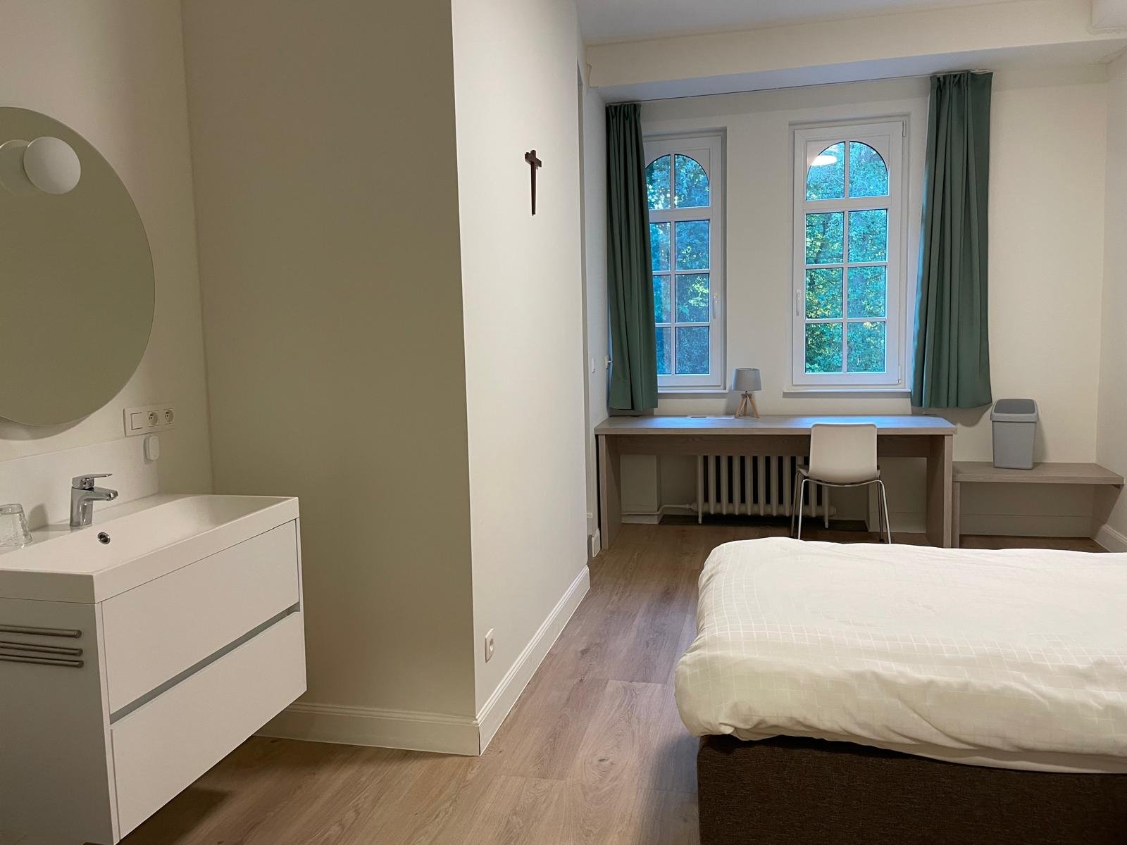 Slaapkamer met een bed, bureau onder twee grote ramen, groene gordijnen, een kloklampje, een witte stoel, en een ventilator.
