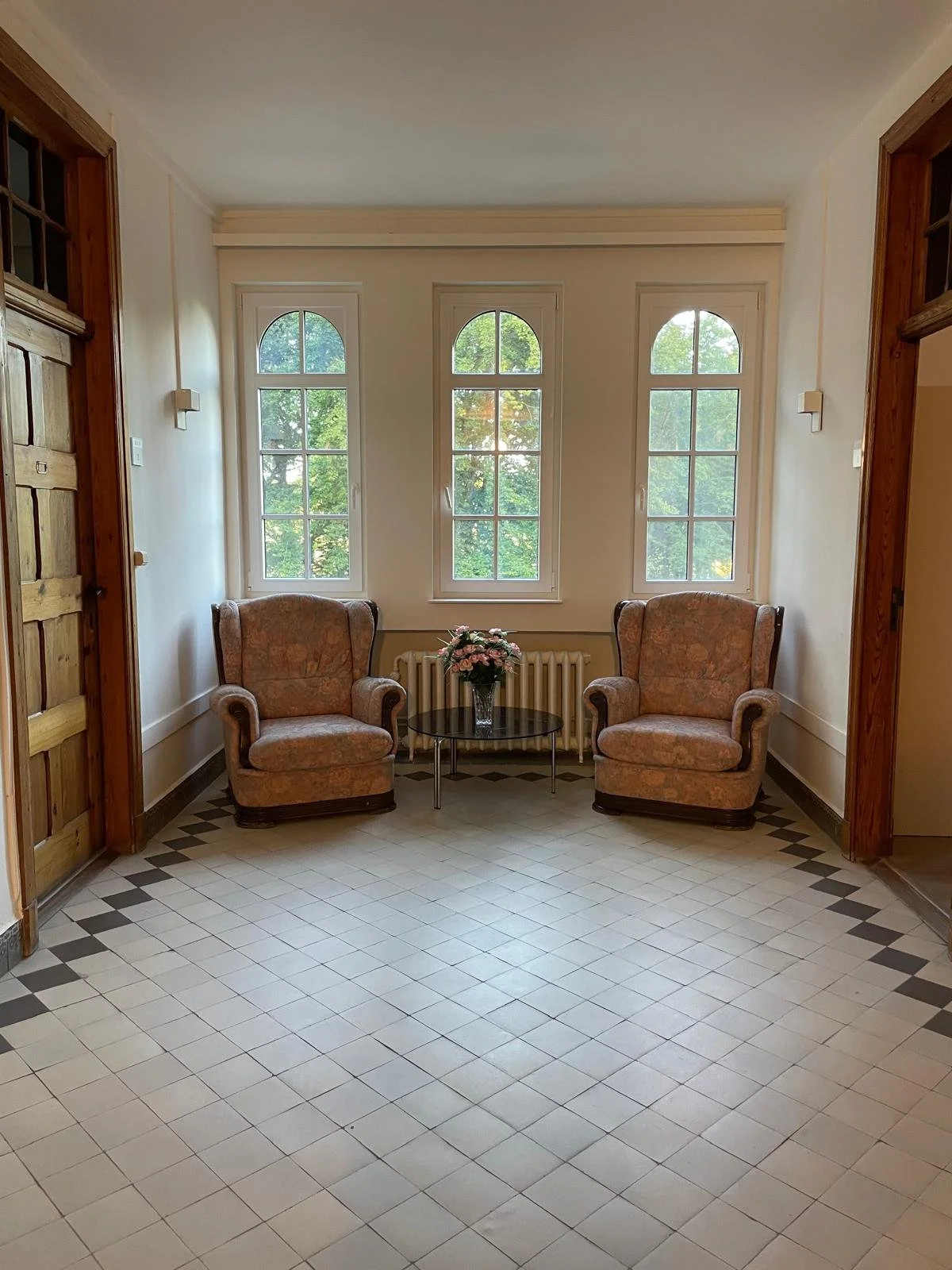 Twee vintage fauteuils met bloemenkransen op een kleine tafel bij grote ramen in een lichte kamer.