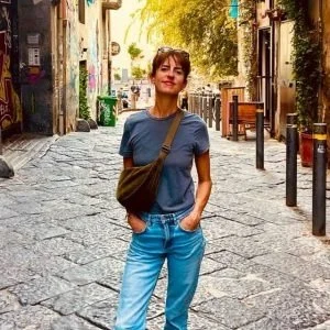 Vrouw staat op een rustige straat met oude gebouwen en bomen, draagt een blauwe T-shirt, lichtblauwe jeans en heeft een schoudertas.