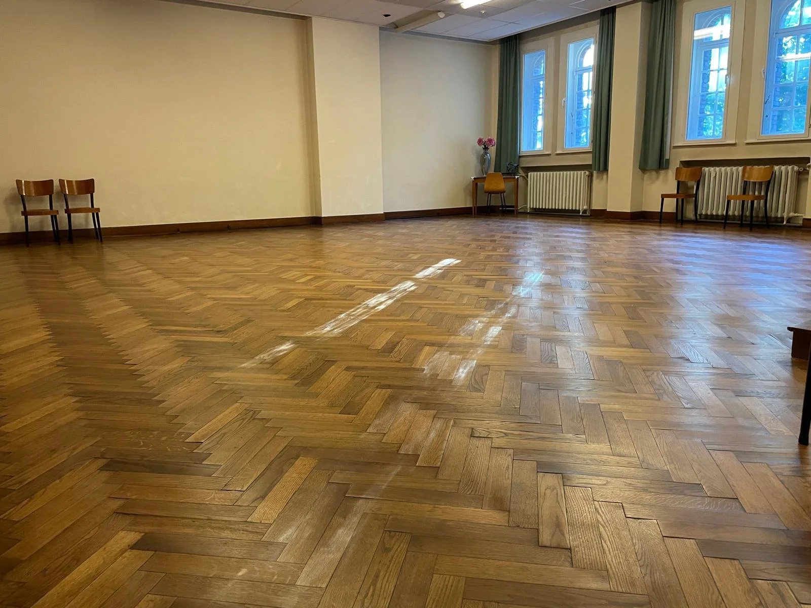 Een lege kamer met houten vloer, drie houten stoelen tegen de muur links, een kleine tafel met vaas en bloemen in de hoek bij het raam. Grote ramen met groene gordijnen.