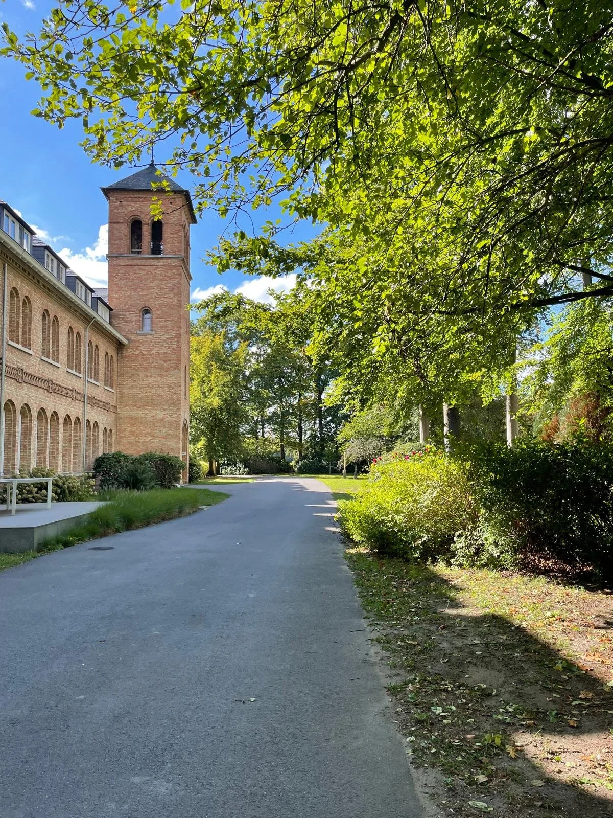 Een pad dat langs een kerk met een toren loopt, omgeven door groene bomen en struiken, onder een blauwe hemel met enkele wolken.