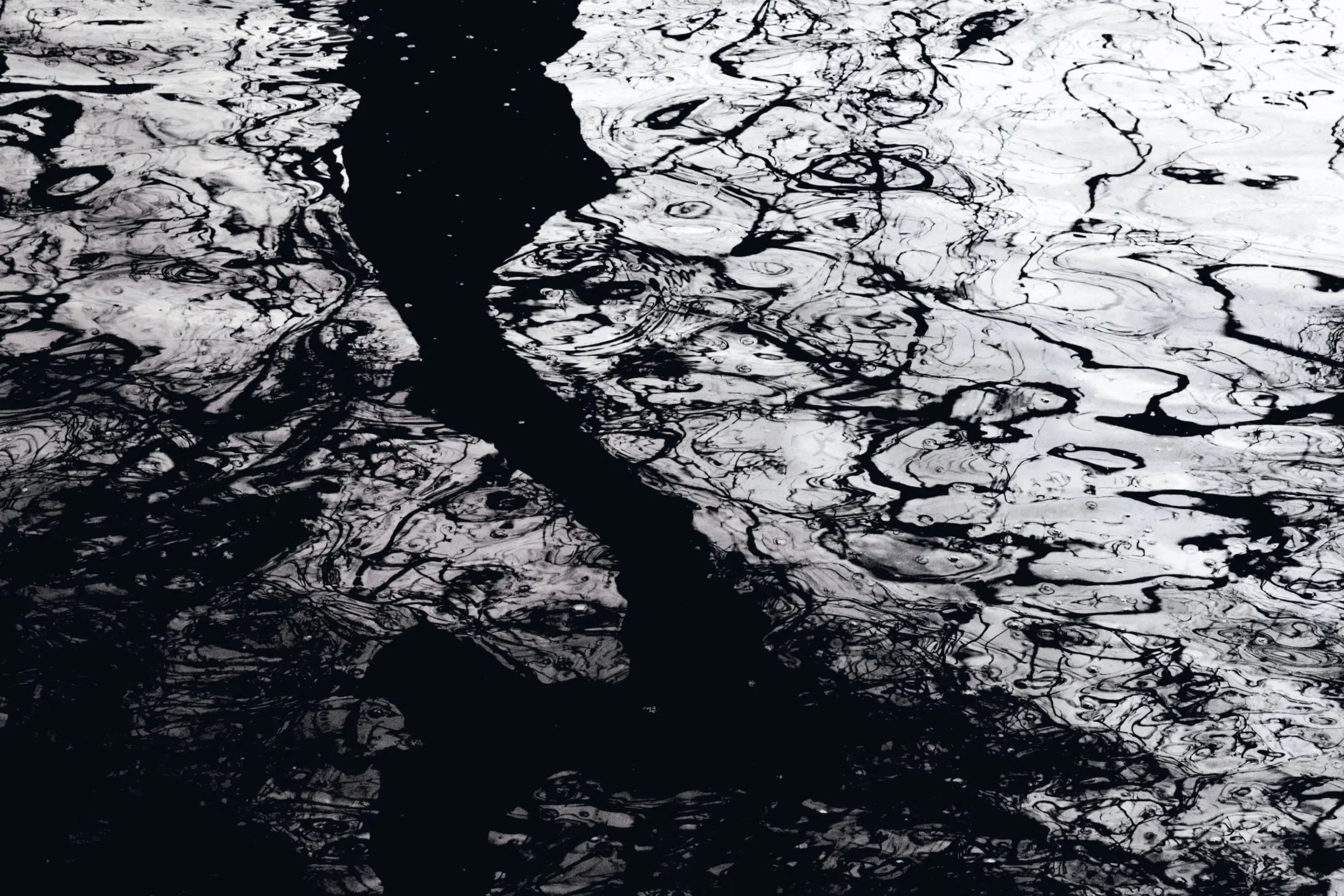 Black and white photo of swirling reflections of trees and sky on water surface.
