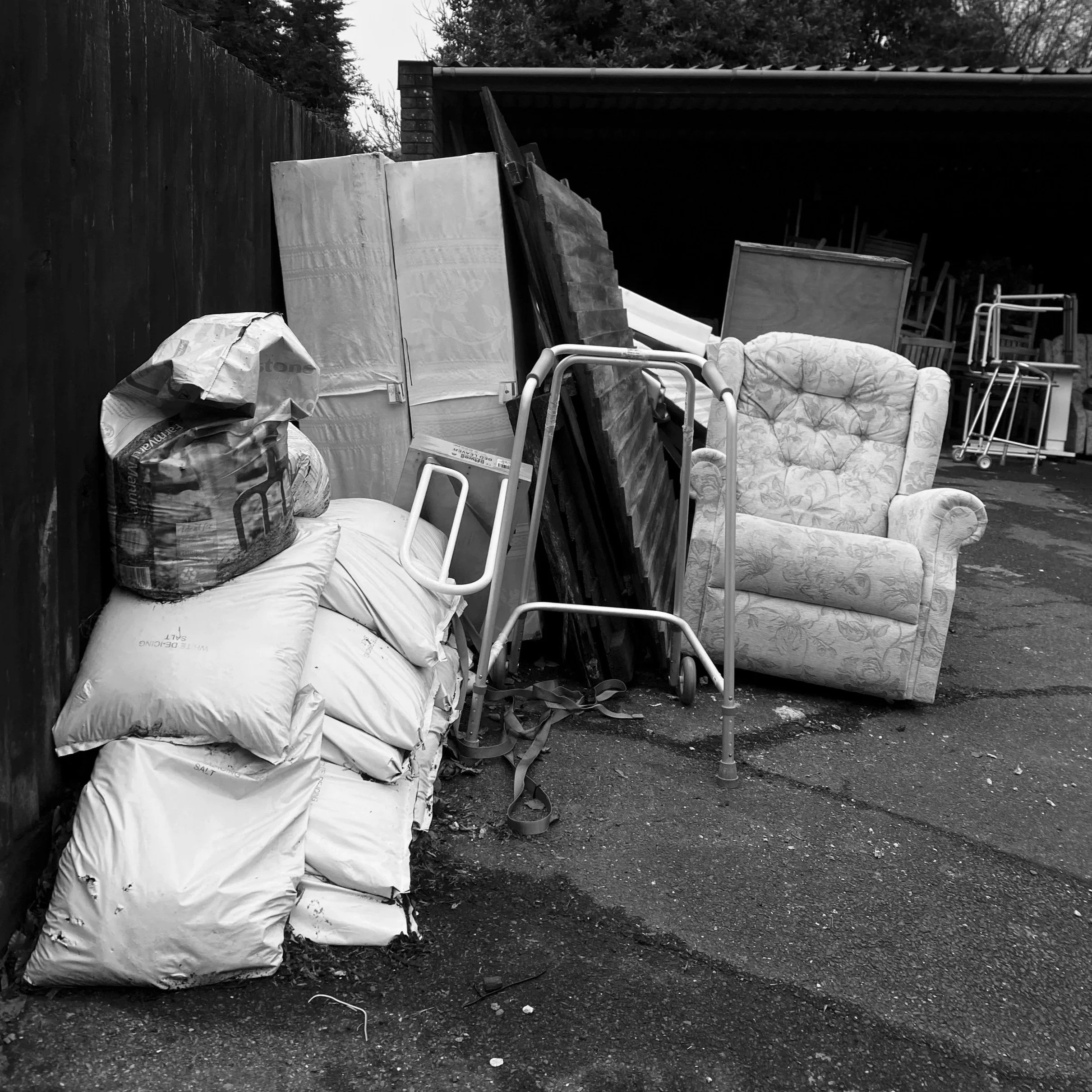 An outdoor scene with bags of trash, furniture, and various discarded items piled against a fence and under a shelter. The ground is dirt and the scene appears cluttered.