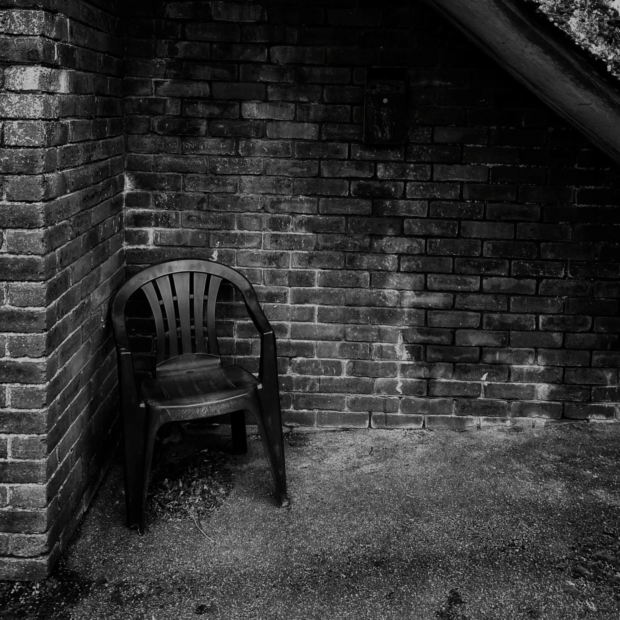 Black and white photo of a corner with brick walls and an umbrella stand with a plastic chair.