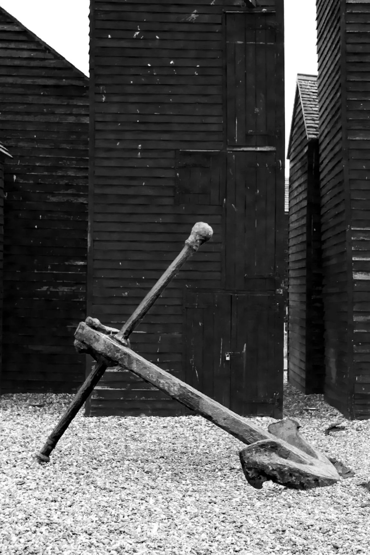 A black and white photograph of a broken wooden plank leaning against another plank, with a gravel ground and tall wooden fences in the background.
