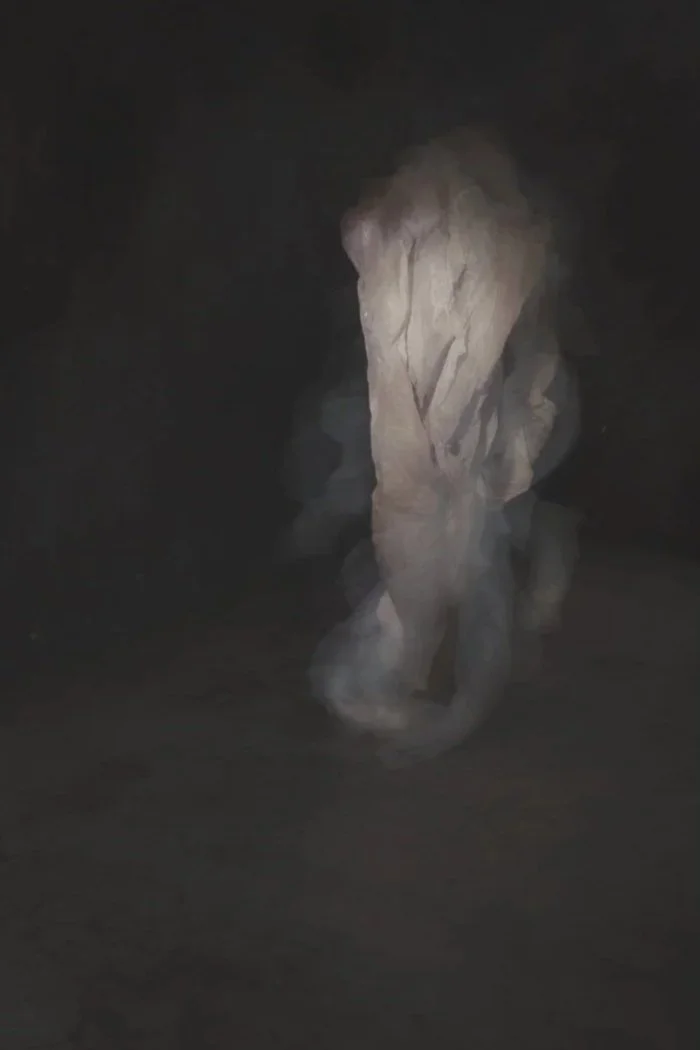 A figure wrapped in a large piece of fabric or paper, standing in a dark space with minimal lighting