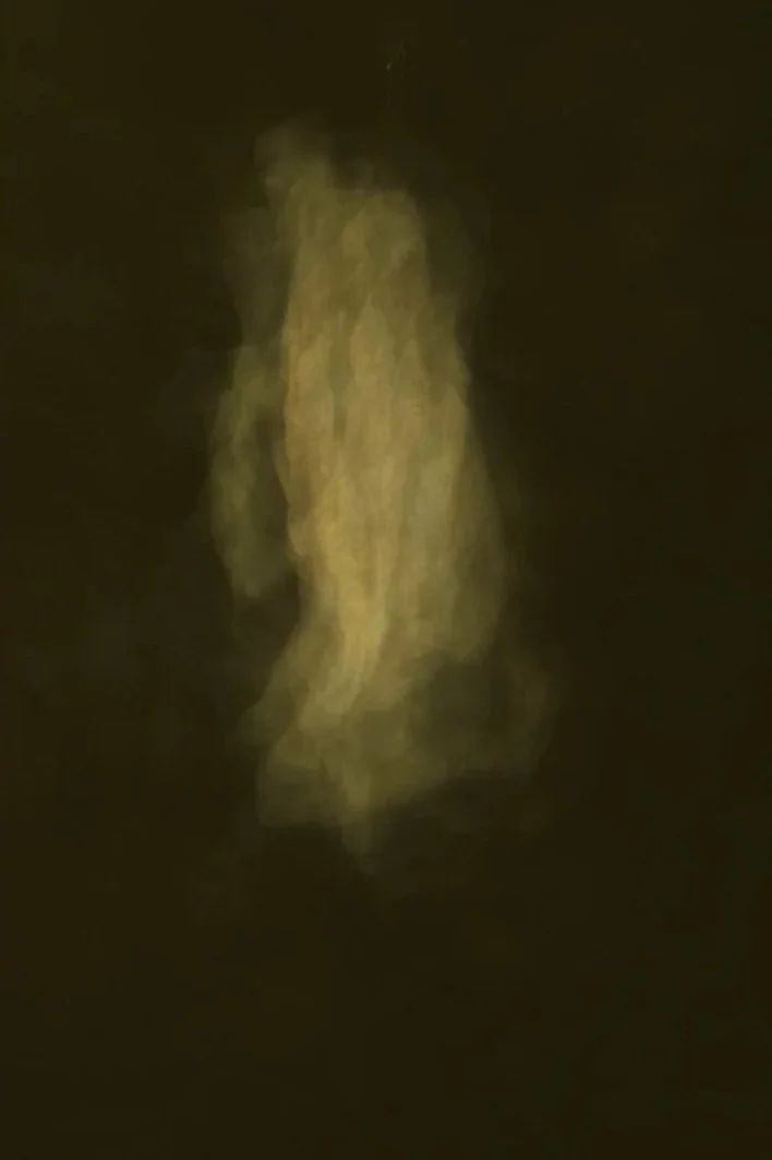 Blurred, yellowish figure standing against a dark background, with indistinct features