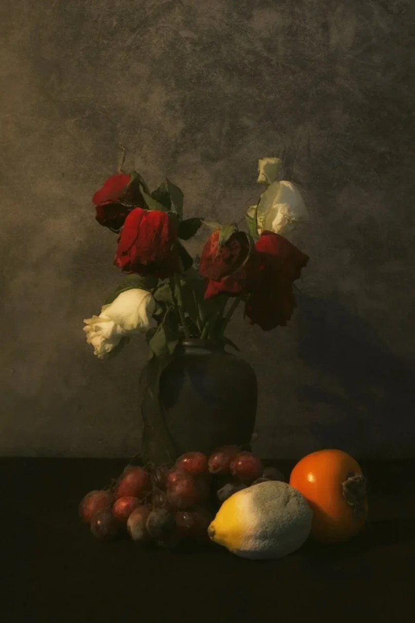 A dark vase holding five wilted roses with white and red blooms, placed on a surface alongside grapes, a lemon, and an orange against a textured background.