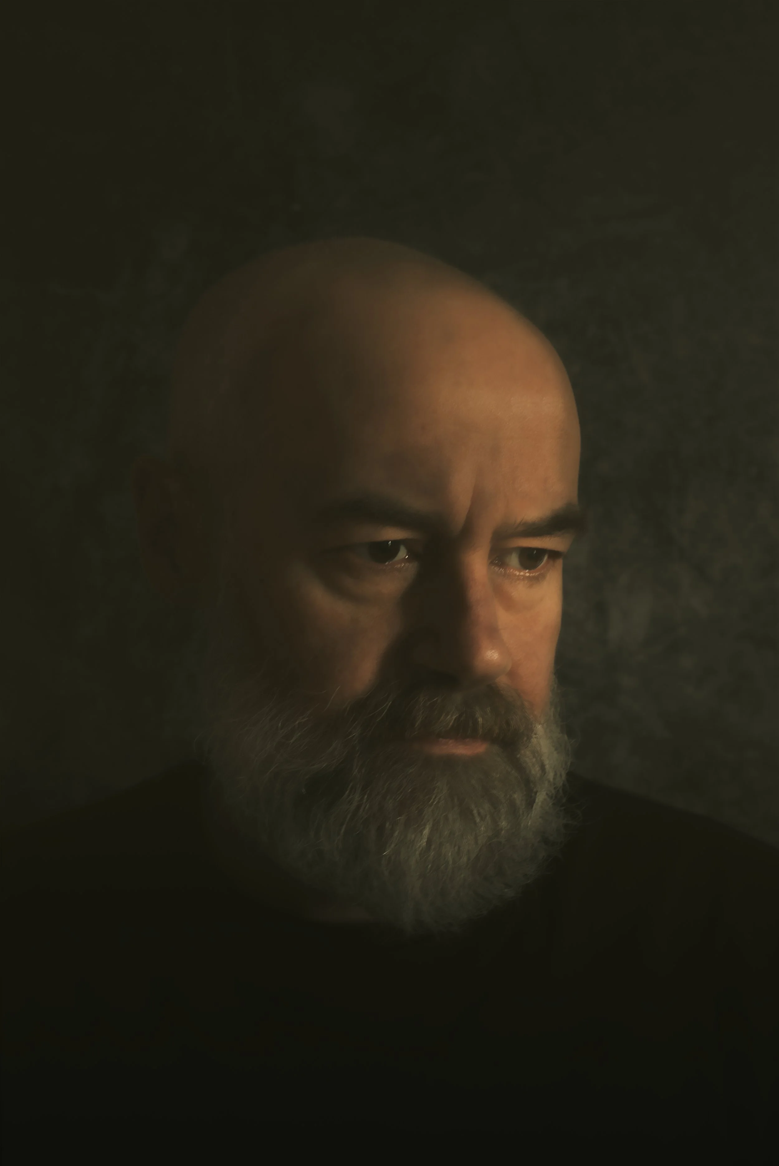 Close-up of a middle-aged man with a bald head and gray beard, looking down in a dimly lit environment.