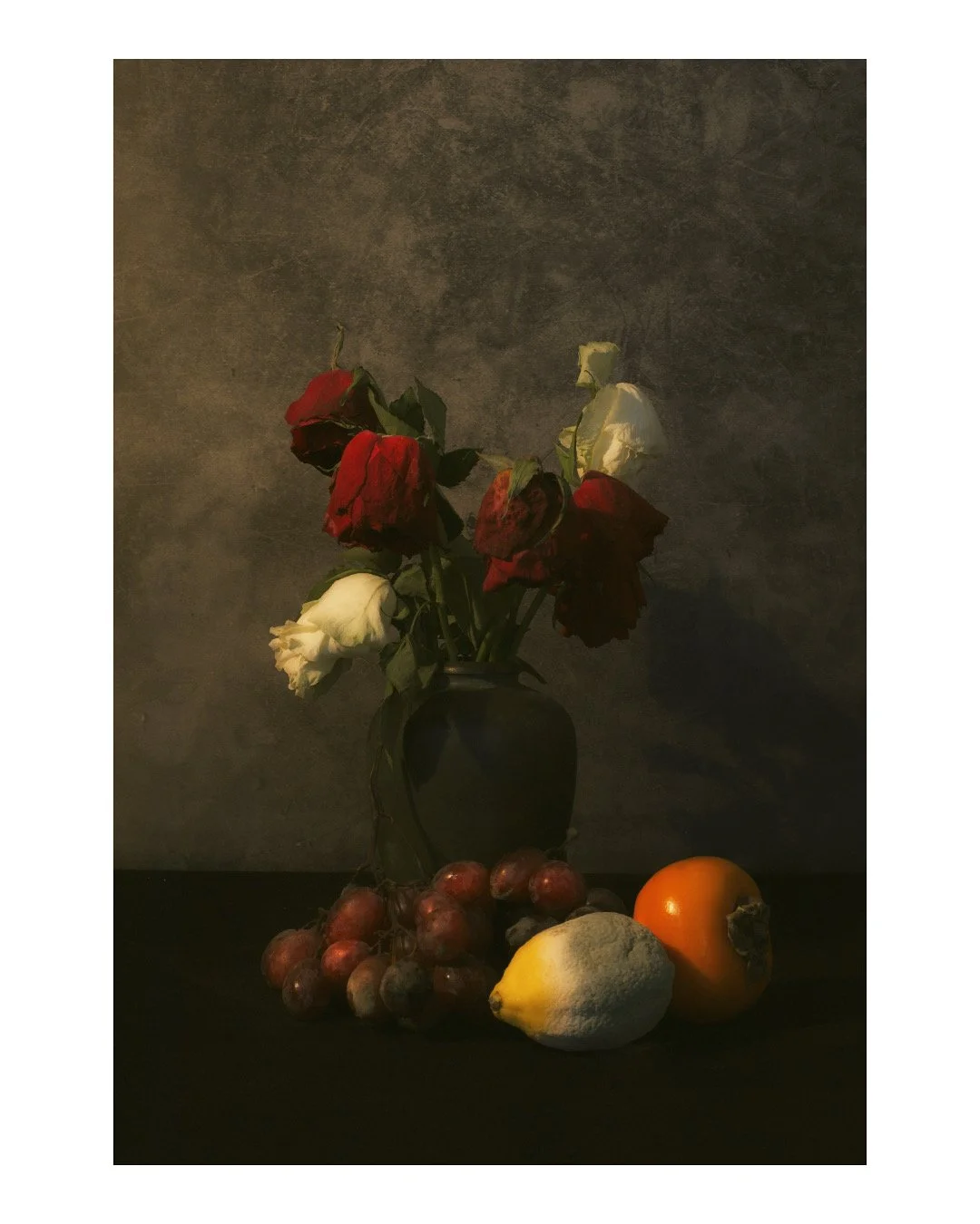 A black vase with wilted red and white roses, surrounded by a bunch of grapes, a lemon, and a persimmon on a dark surface against a textured gray background.