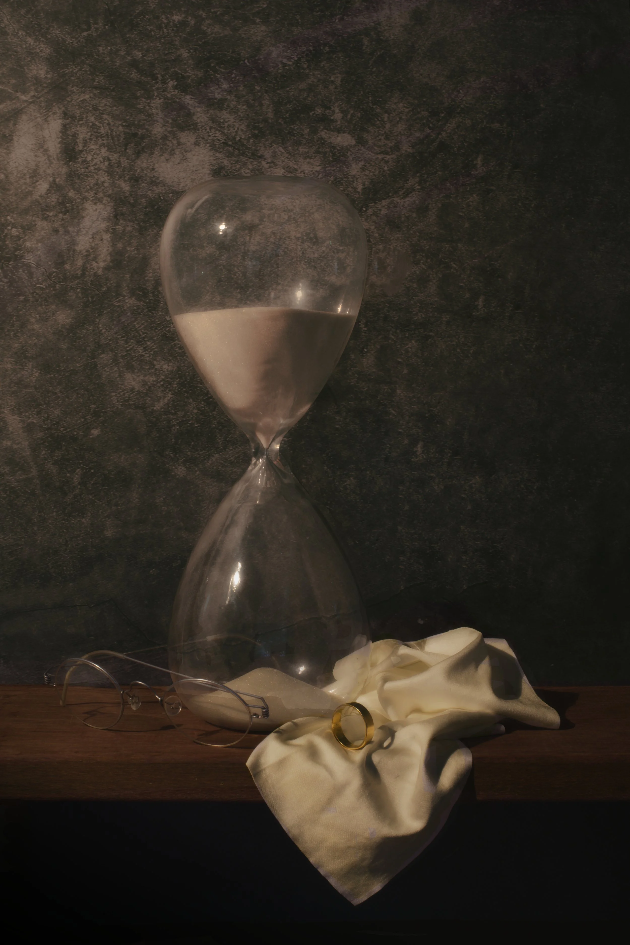 A glass hourglass filled with white sand on a wooden surface, surrounded by a pair of glasses, a white cloth, and gold rings, against a dark textured background.