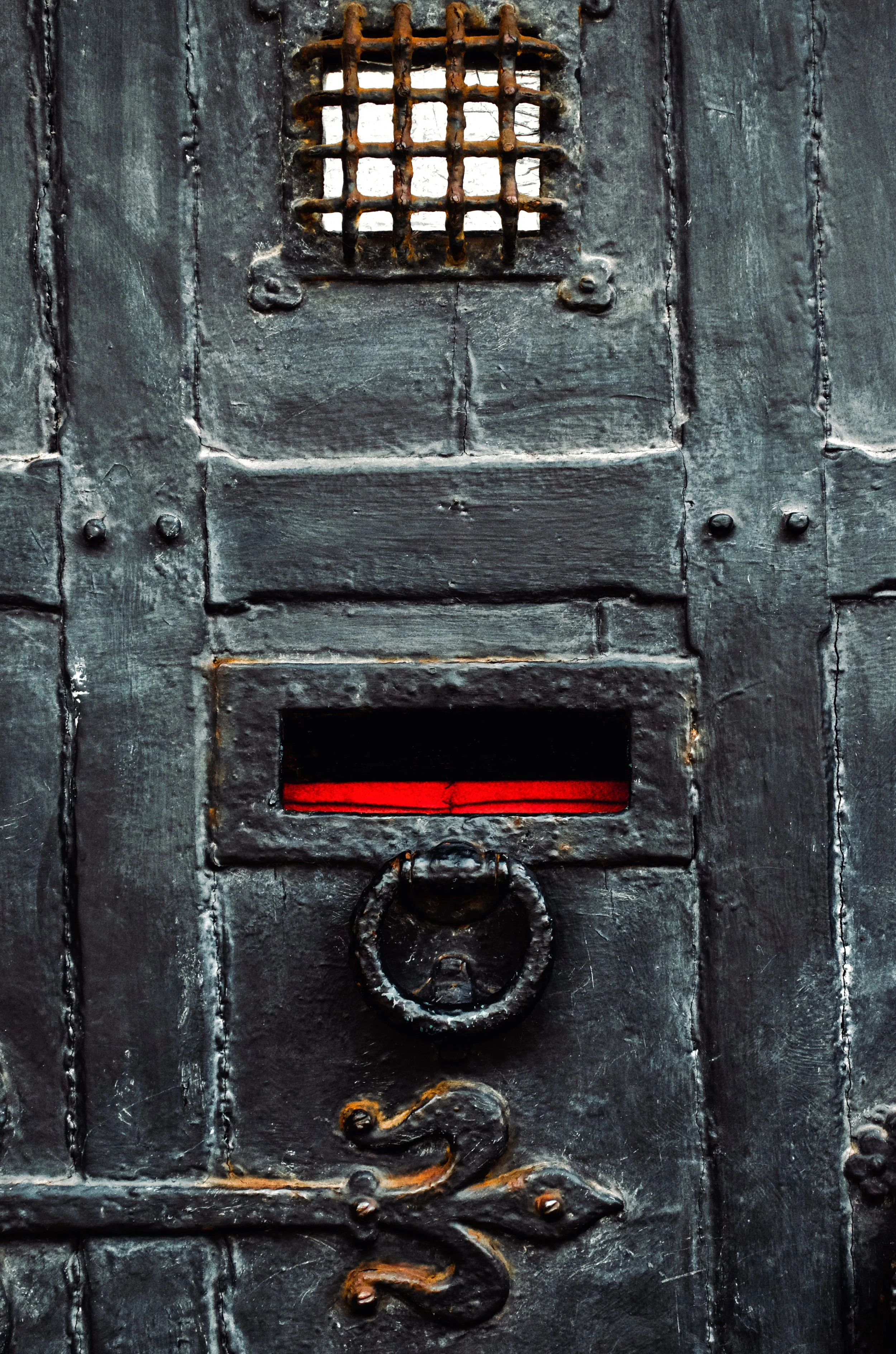 Close-up of an old, black metal door with a small barred window, a red horizontal opening, and a decorative ring handle.