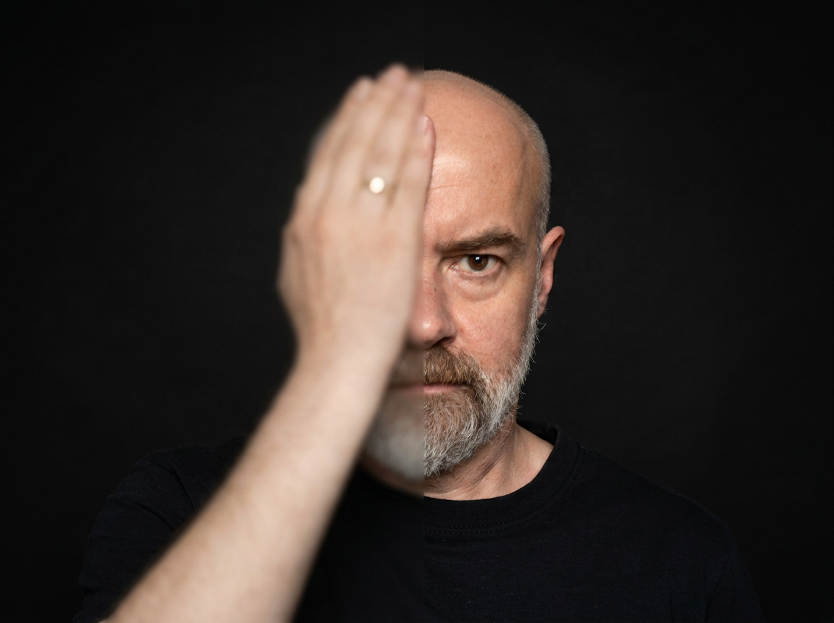 A man with a beard and bald head looks directly at the camera with his face partially obscured by his raised hand against a black background.