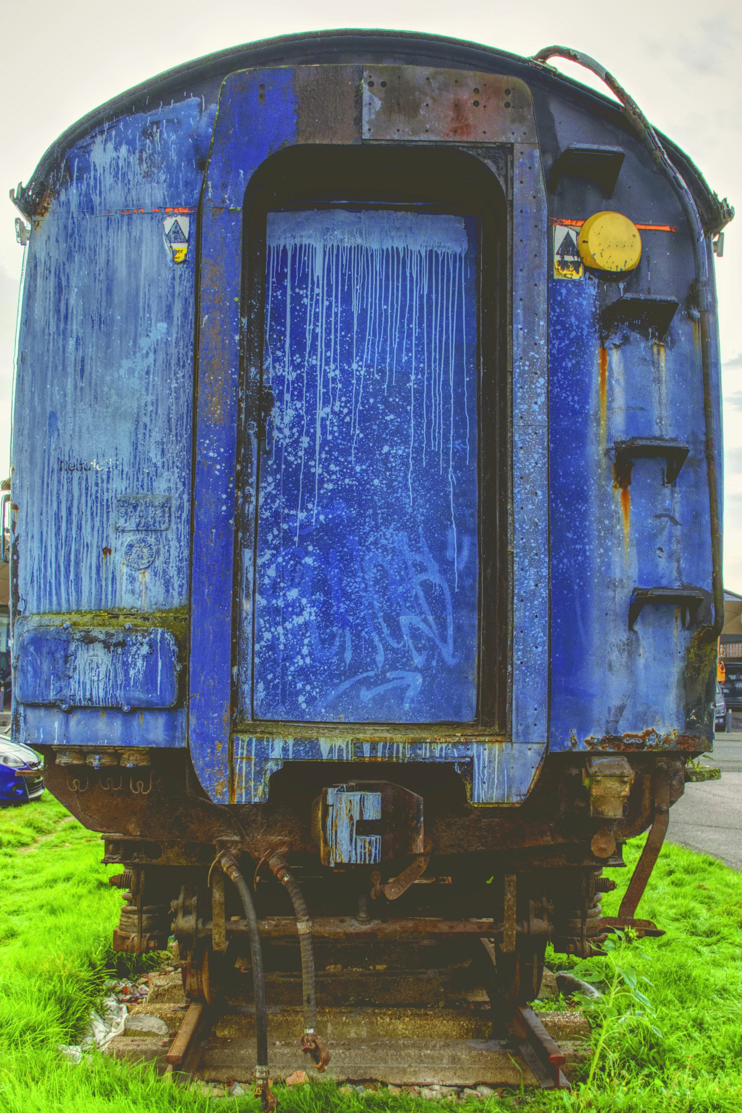 An old, rusted train car with peeling blue paint, sitting on tracks, surrounded by green grass and small plants.