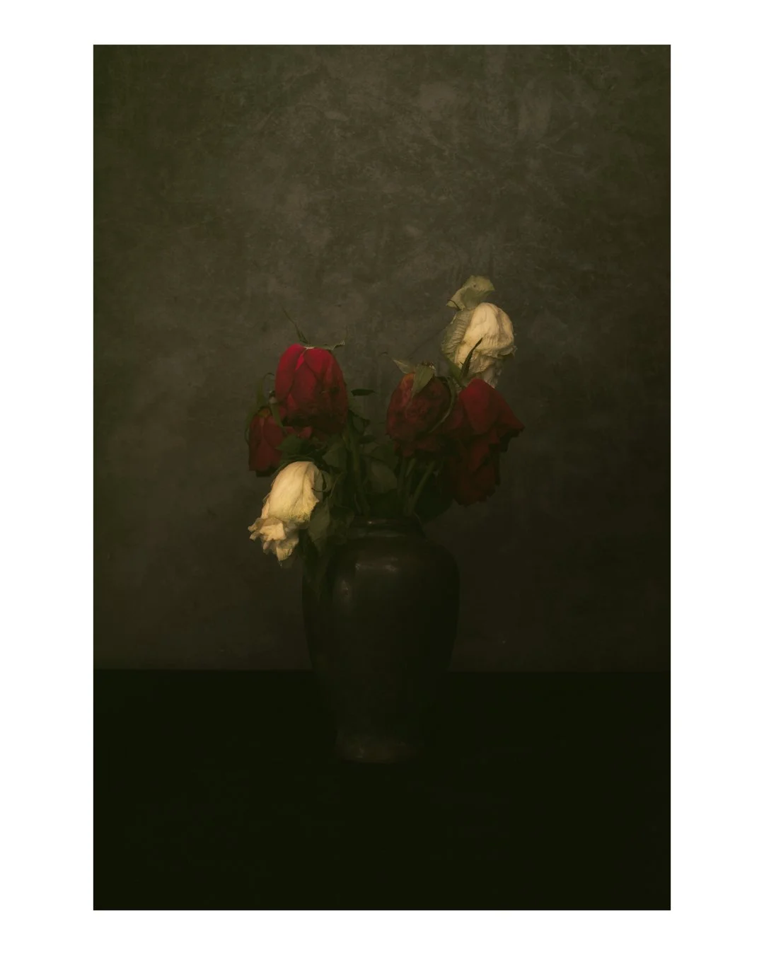 A dark vase holding wilted red and white roses against a dark background.