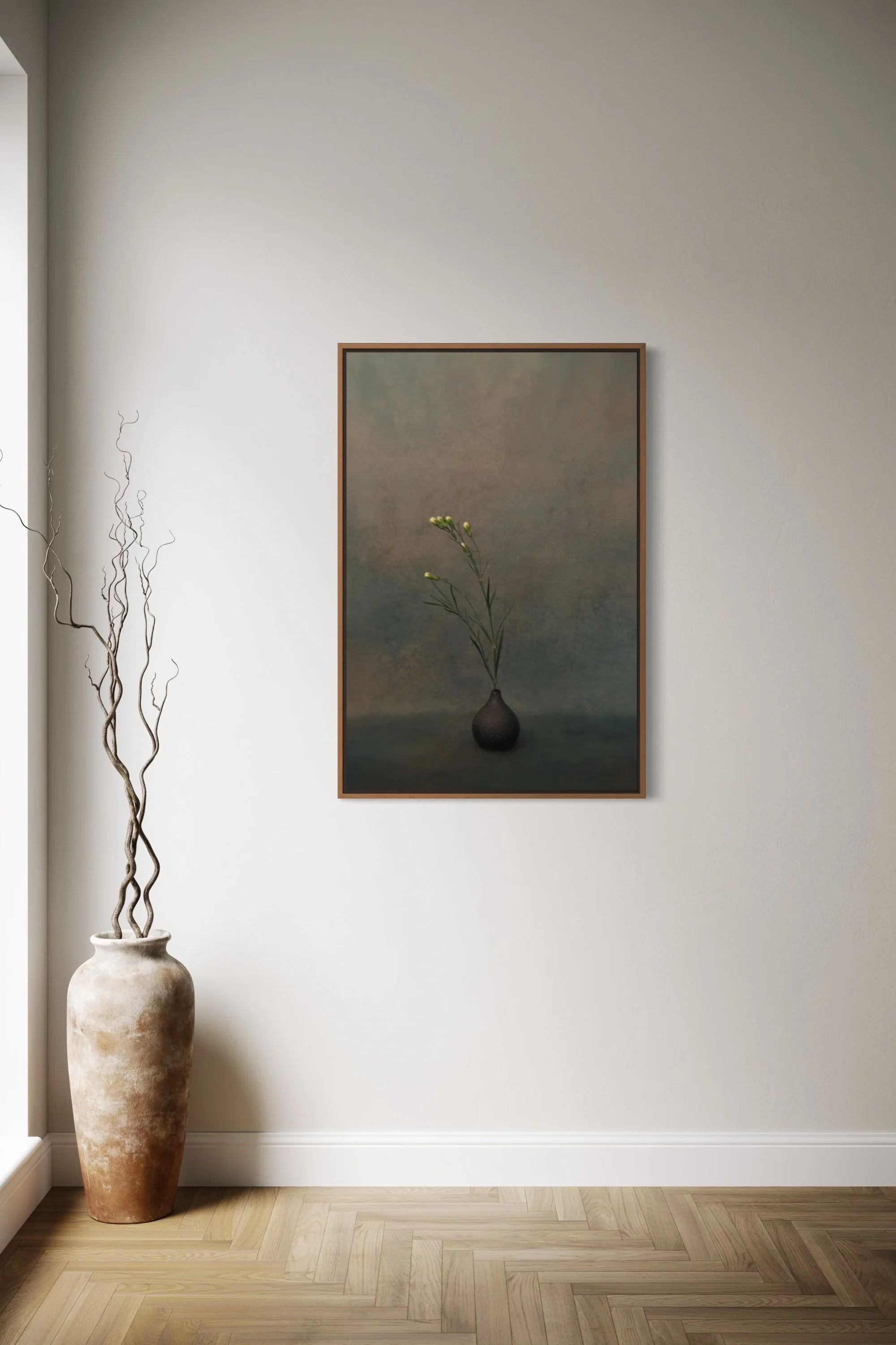 A tall, beige, weathered ceramic vase with two twisting, leafless, decorative branches stands on the floor in front of a plain white wall. To the right of the vase, there's a framed painting of a single green plant with white flowers in a dark vase, hanging on the wall. The floor is light-colored hardwood arranged in a herringbone pattern.