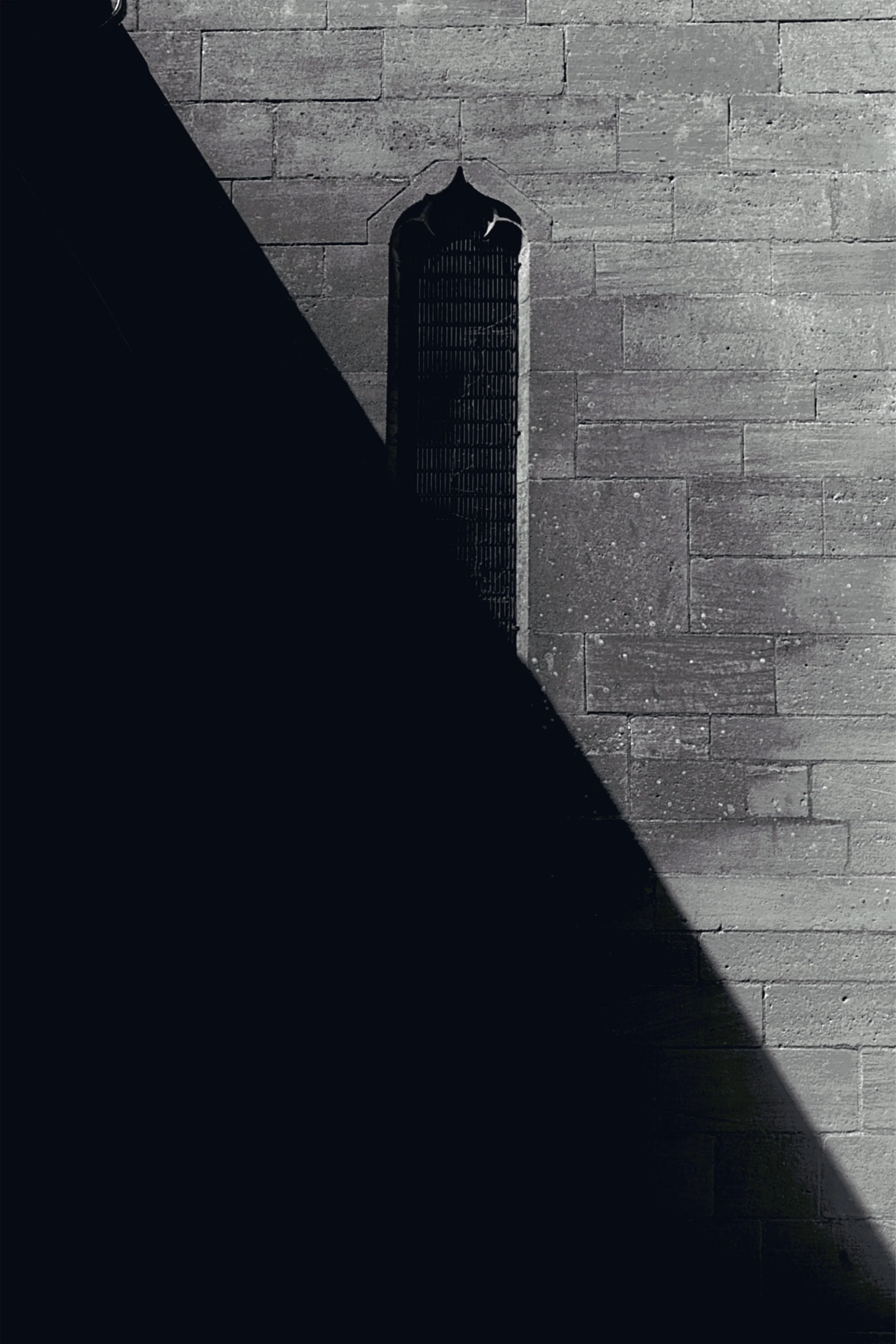 Black and white photo of a stone wall and a narrow, gothic-style window with bars, partially covered by a large shadow.