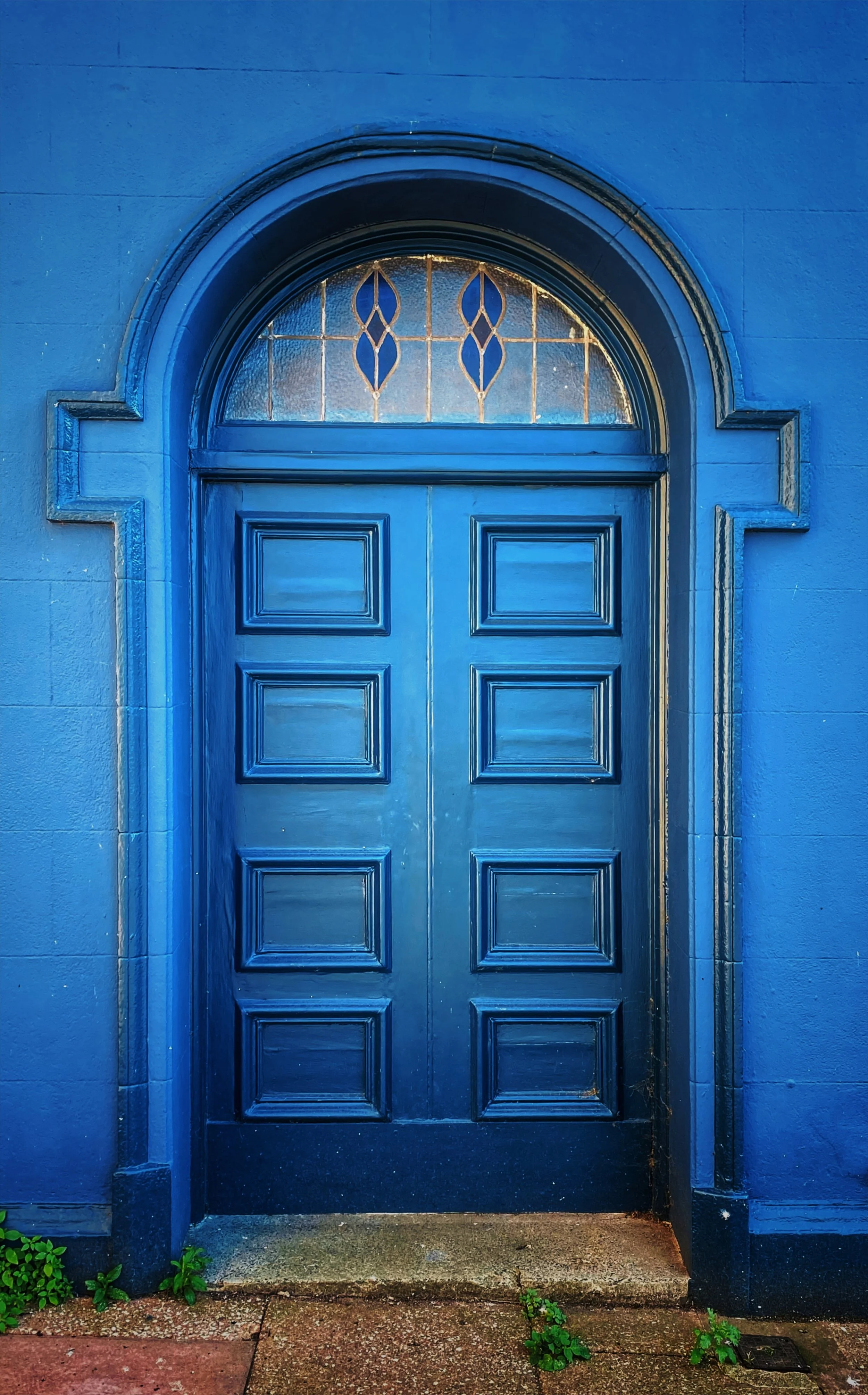 Blue wooden door with decorative window above, set in a blue wall with stone trim, small green plants growing at the base, wet sidewalk in front.