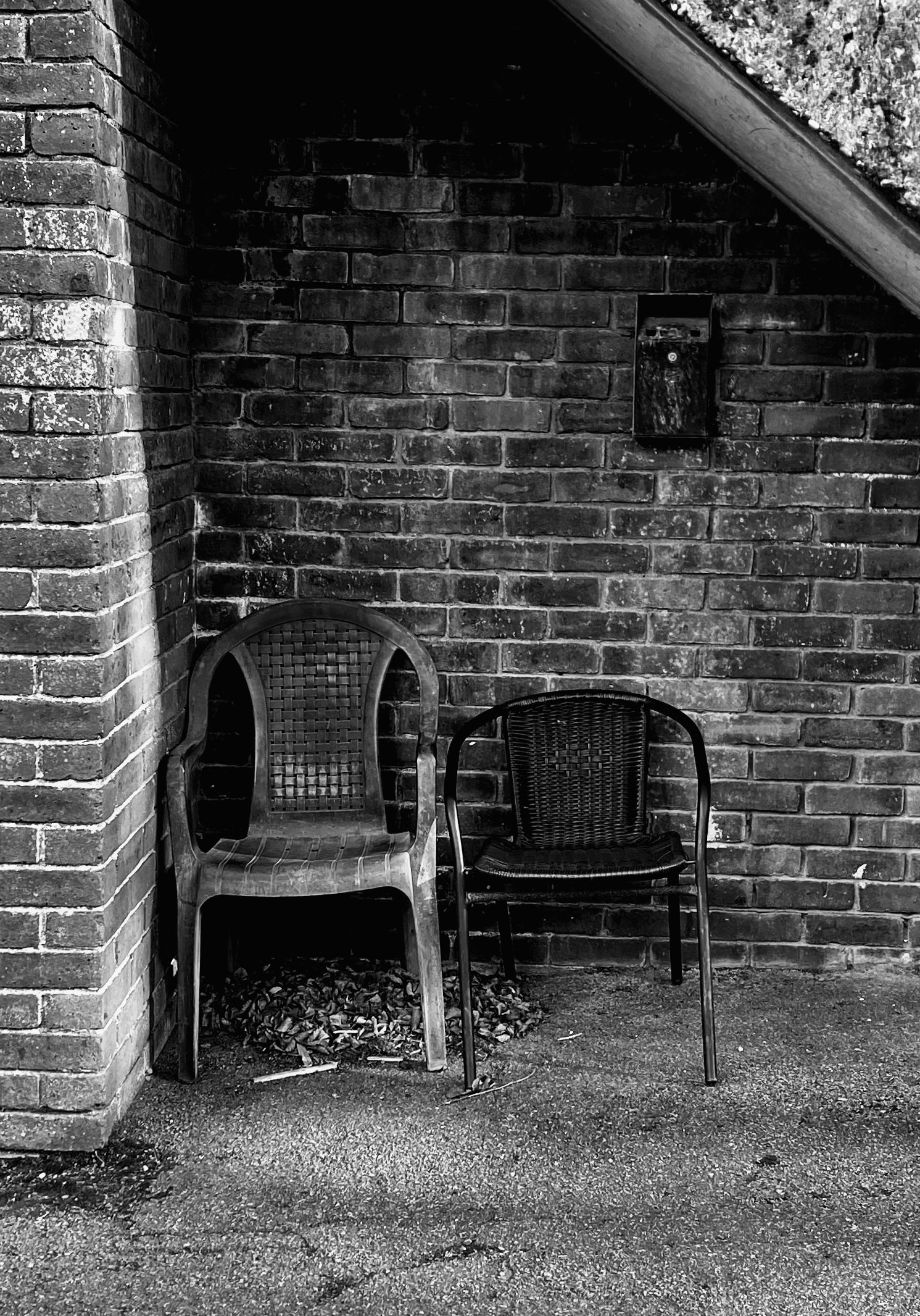 Two chairs, one made of plastic and the other of metal, are placed against a brick wall beneath a stairwell. There is a pile of leaves on the ground under the chairs.