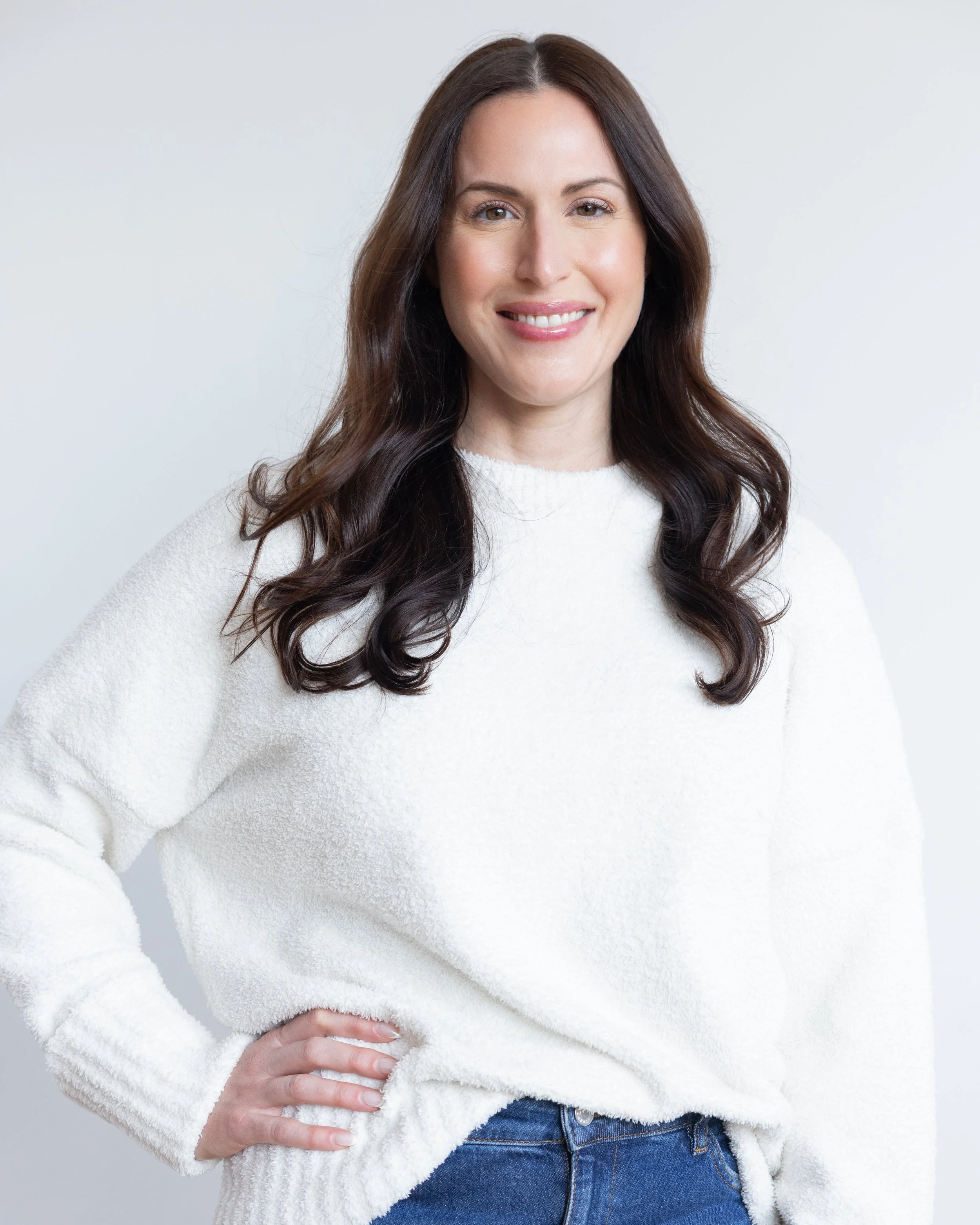 Close-up of a woman with long brown hair, wearing a white fuzzy sweater and jeans, standing against a light gray background.