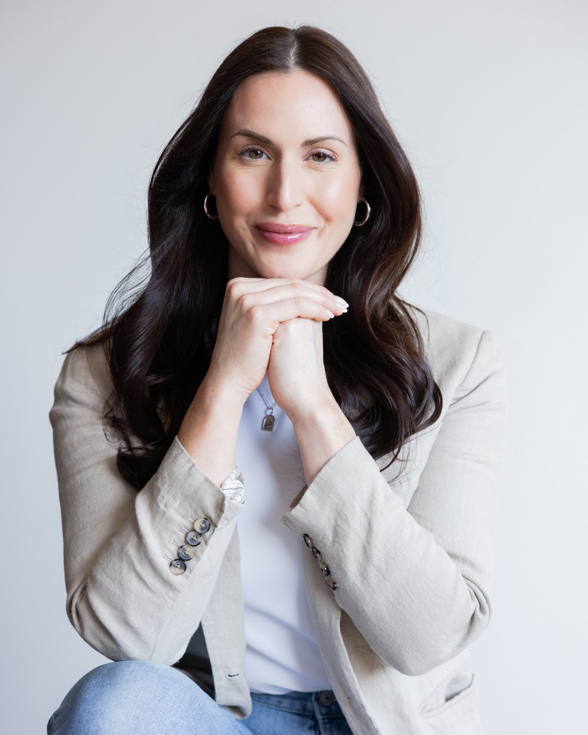 A young woman with long dark hair and hoop earrings, smiling softly with hands clasped under her chin, wearing a beige blazer and light blue jeans, sitting against a plain white background.