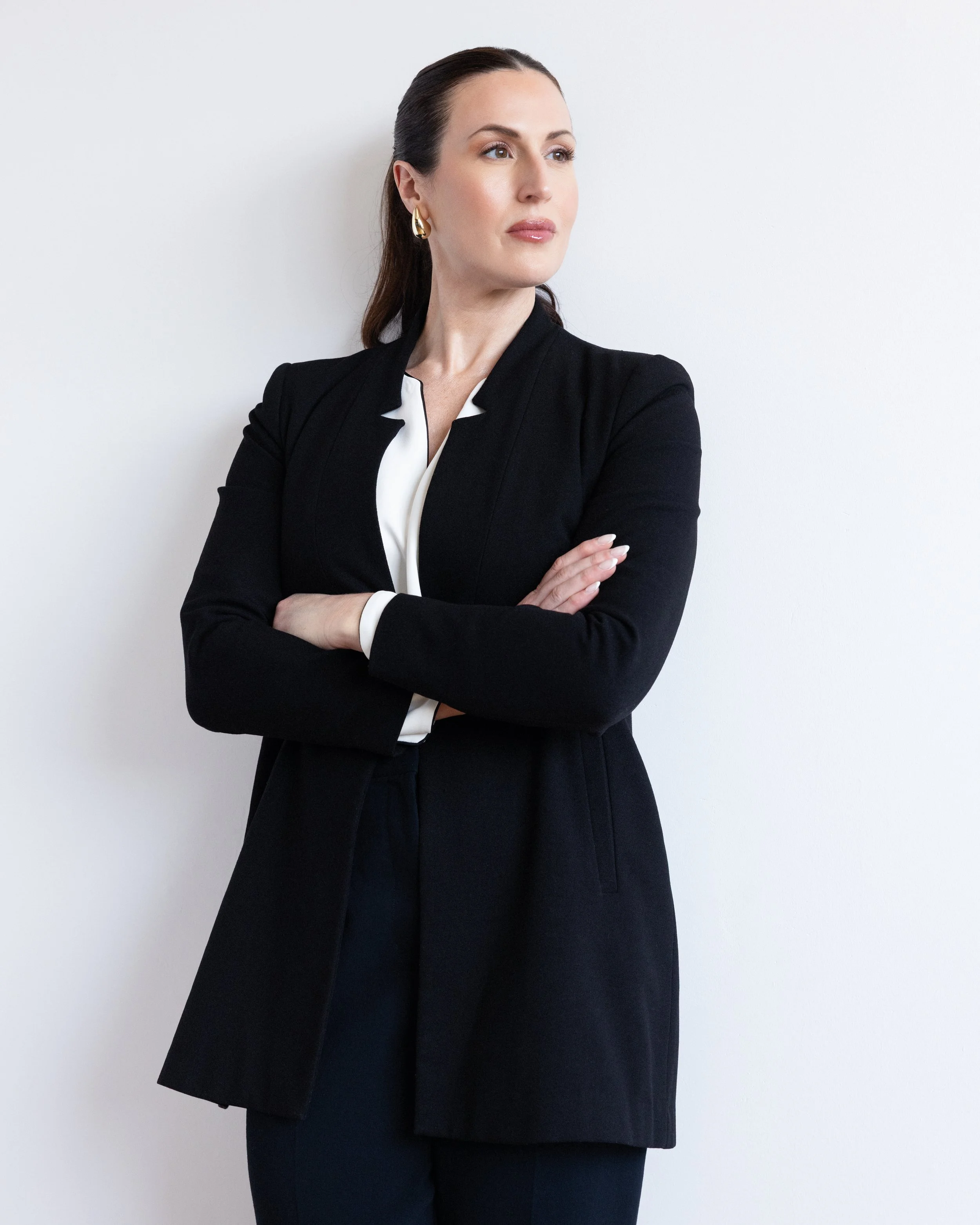 A woman with dark brown hair pulled back, wearing a black blazer, white blouse, gold hoop earrings, and light makeup, stands with her arms crossed against a plain white wall.