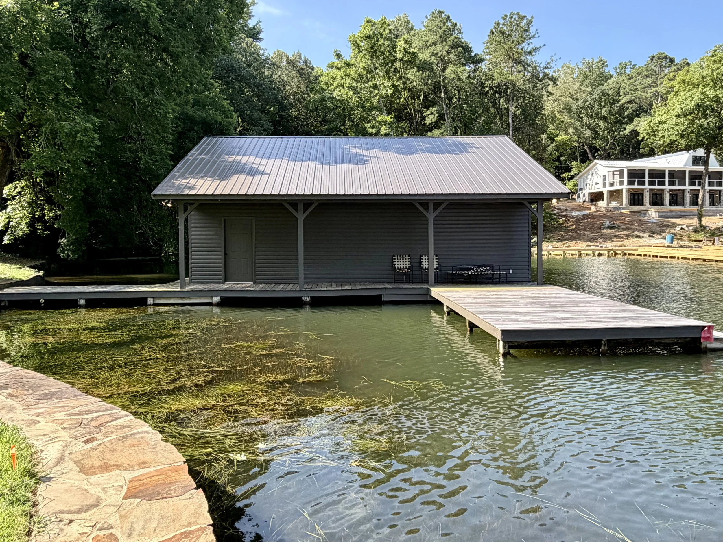 A lakeside scene with a gray building on stilts, a dock extending over water, and trees in the background under a clear blue sky.
