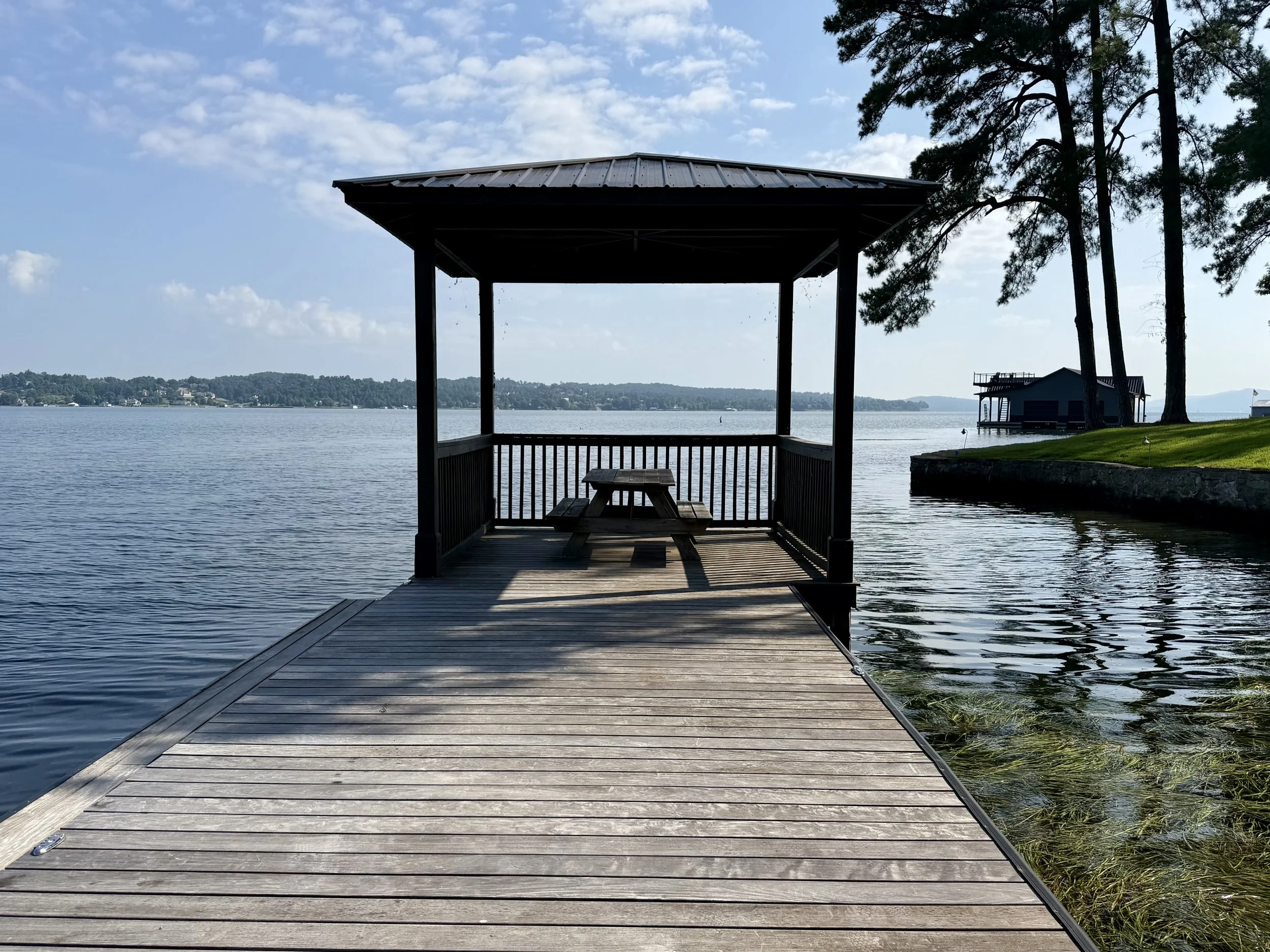 A wooden dock extending over water, with a small gazebo on the end, overlooking a lake with a house nearby and trees on the shoreline.
