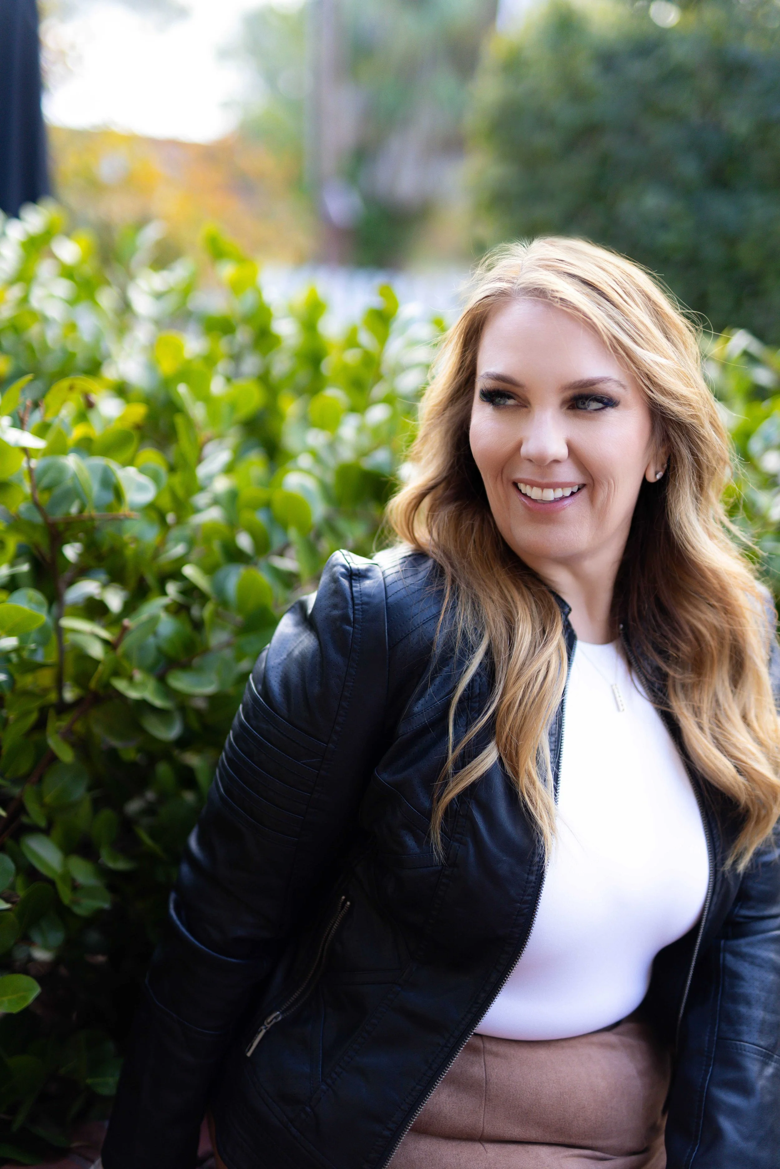 A woman with wavy blonde hair smiling, wearing a black leather jacket and white shirt outdoors next to green foliage.