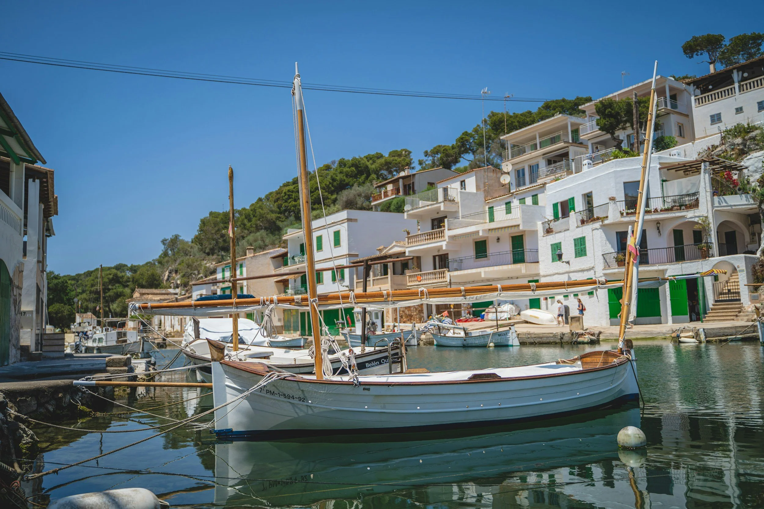 A small harbour with boats docked along the water and white buildings with green shutters on a hillside under a clear blue sky.